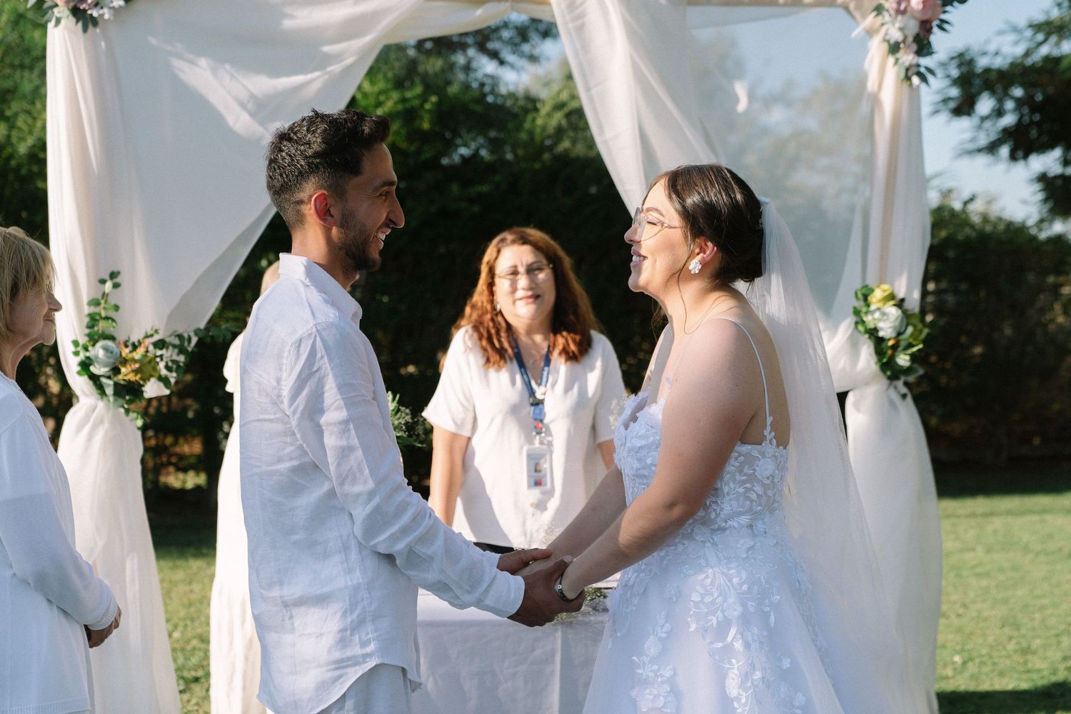 Bride and groom hold hands smiling during outdoor wedding ceremony under white floral arch with officiant.