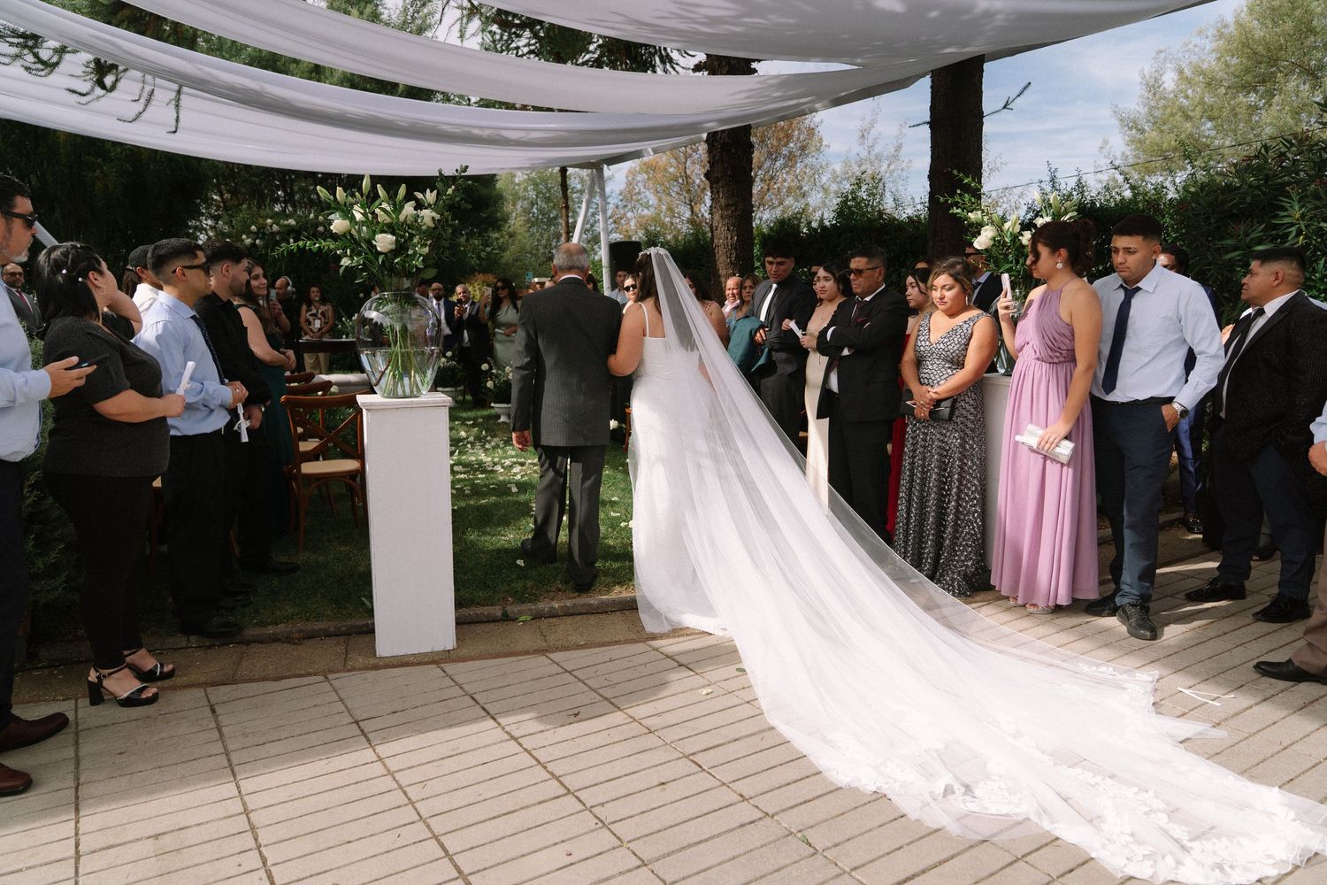 Outdoor wedding ceremony under a white canopy with bride in long veil and guests gathered.