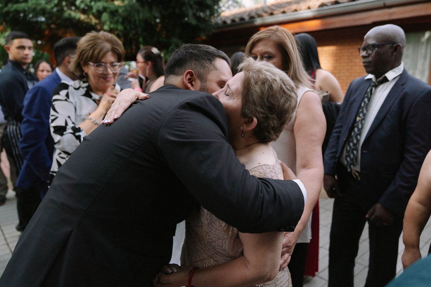 Groom in black suit kisses elderly woman on cheek while guests gather at outdoor wedding reception.
