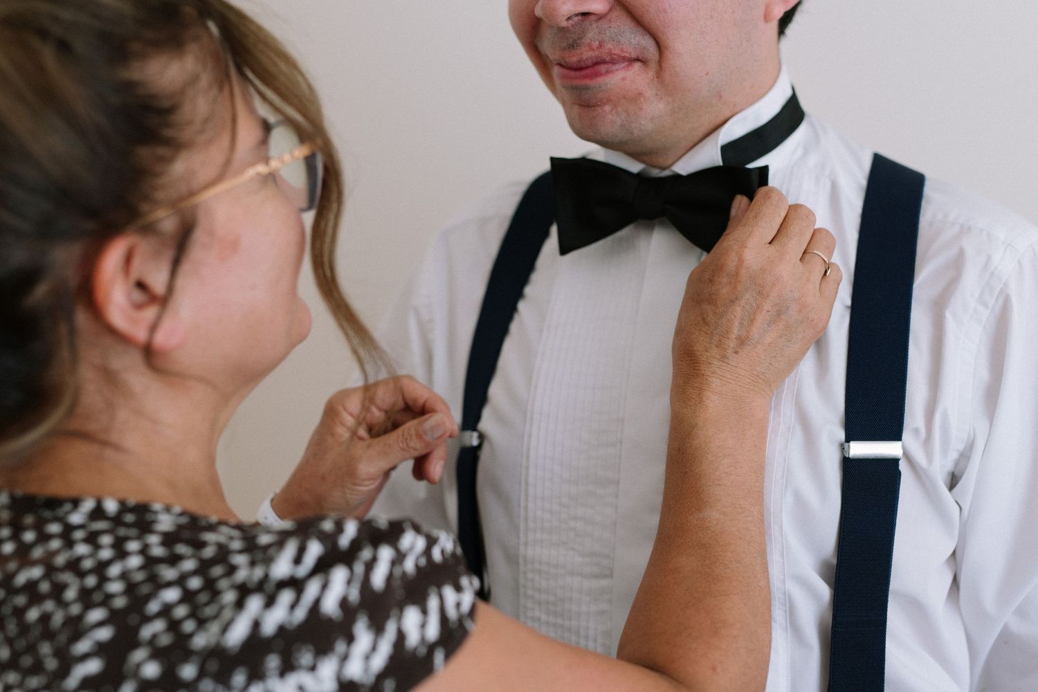 Woman adjusting bow tie on groom wearing white dress shirt and suspenders at formal event.