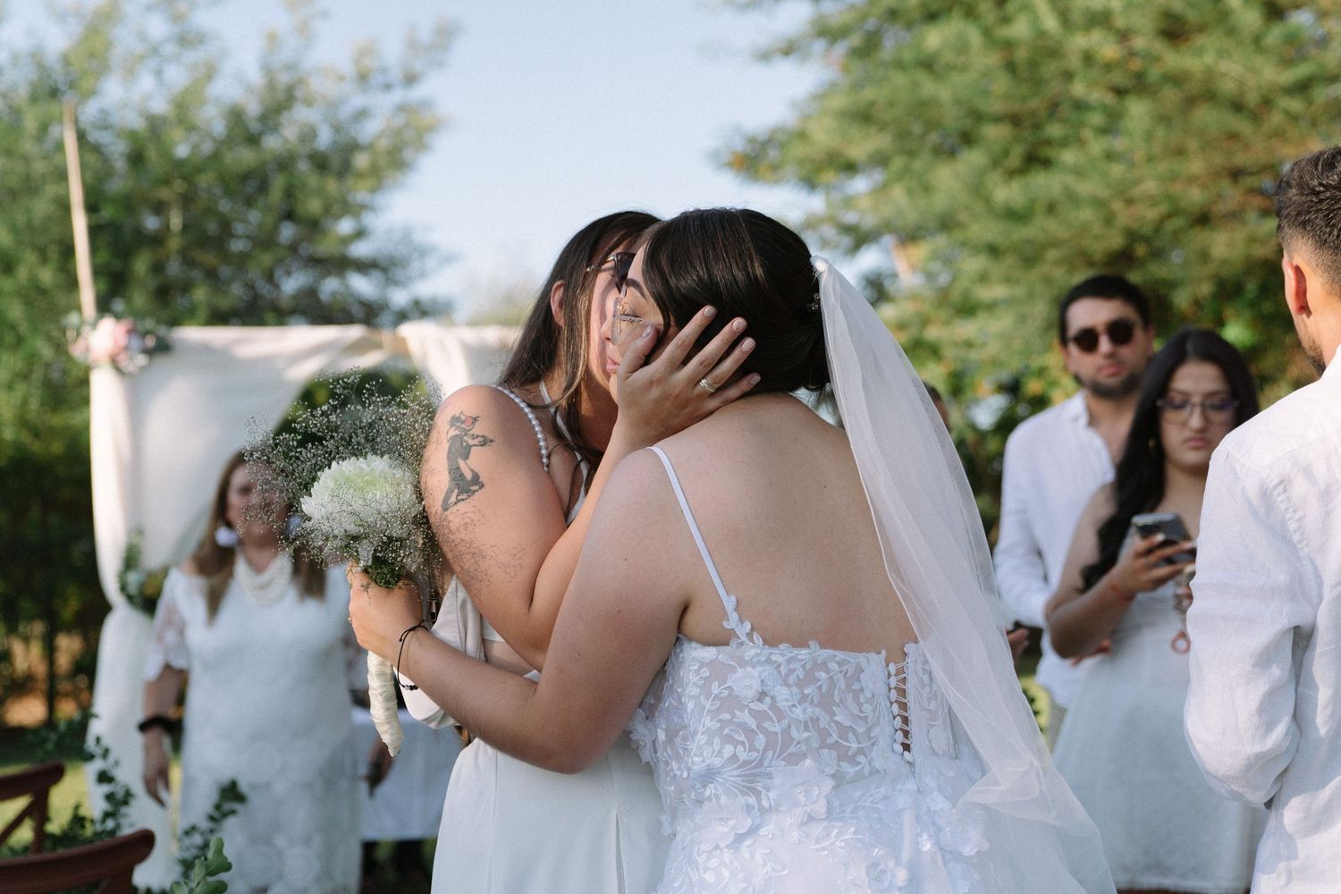 Emotional bride embraces guest in white dress holding bouquet after outdoor wedding ceremony.