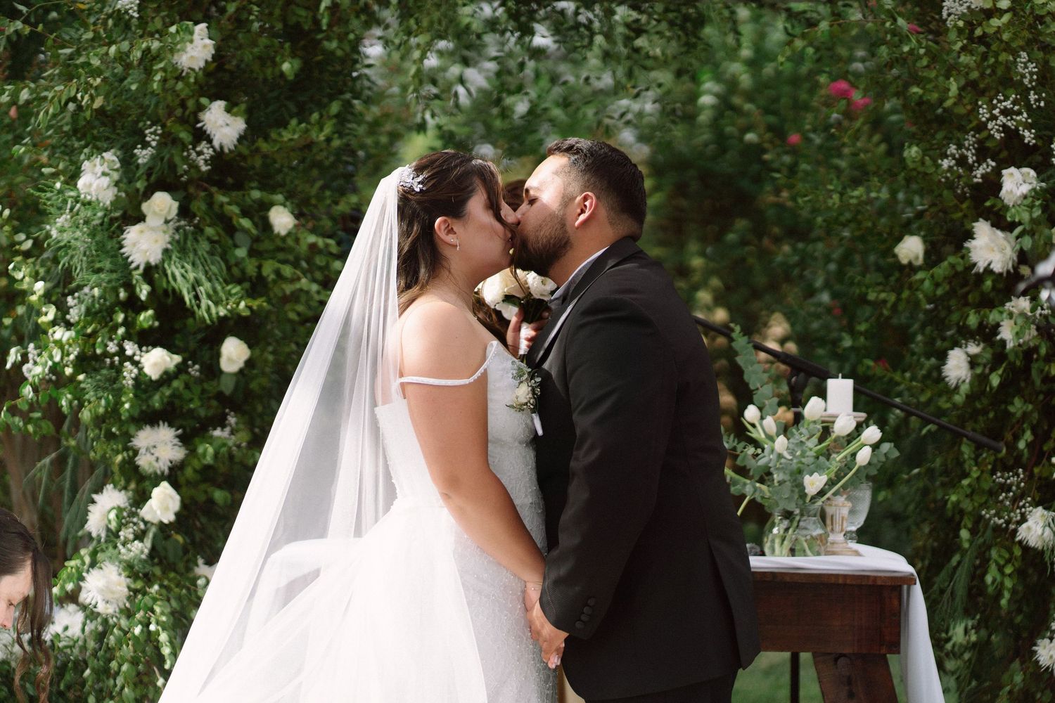 Bride and groom share first kiss at outdoor wedding ceremony surrounded by white floral arch.