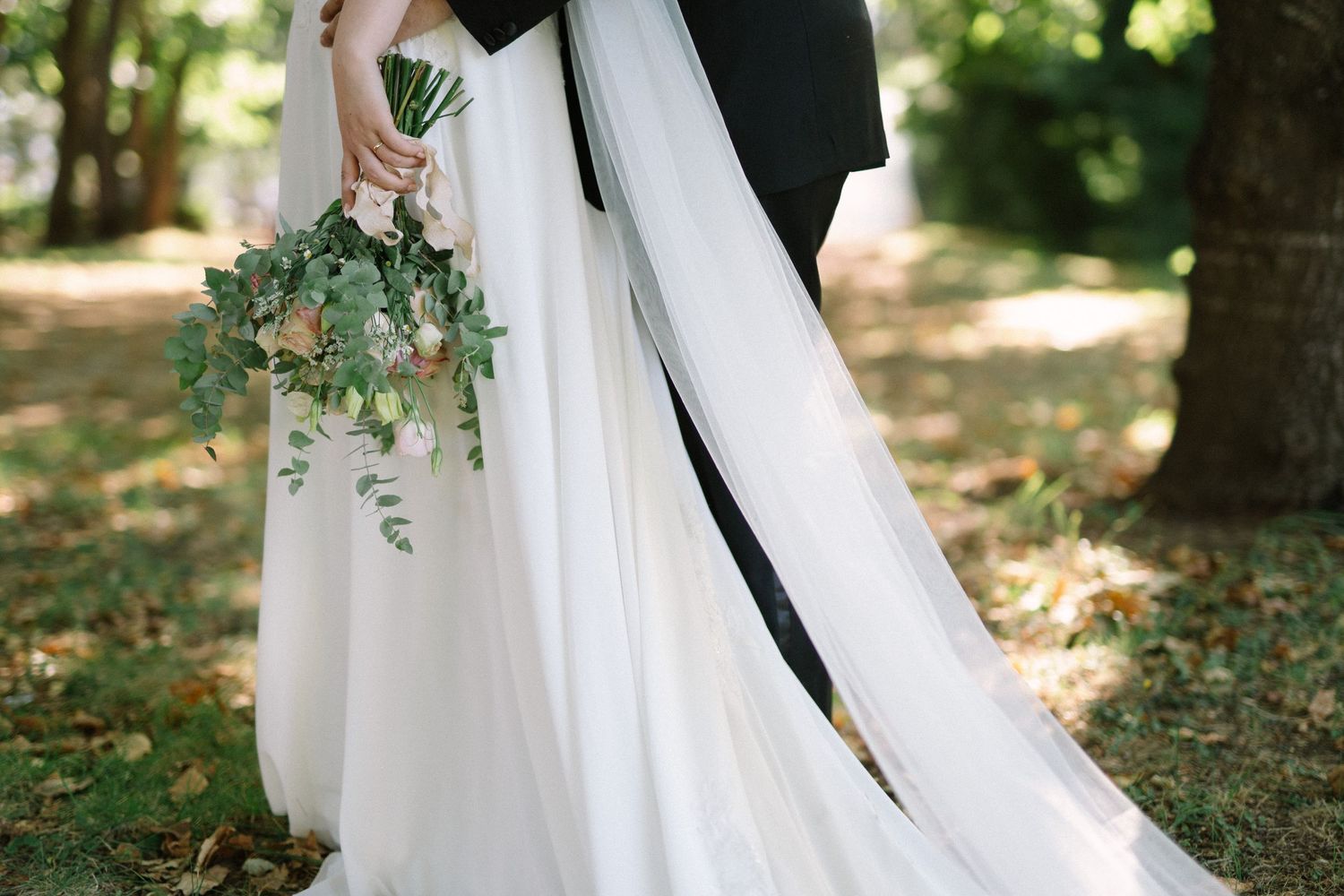 A delicate white wedding dress train flows beside a dark suit in an outdoor garden setting.