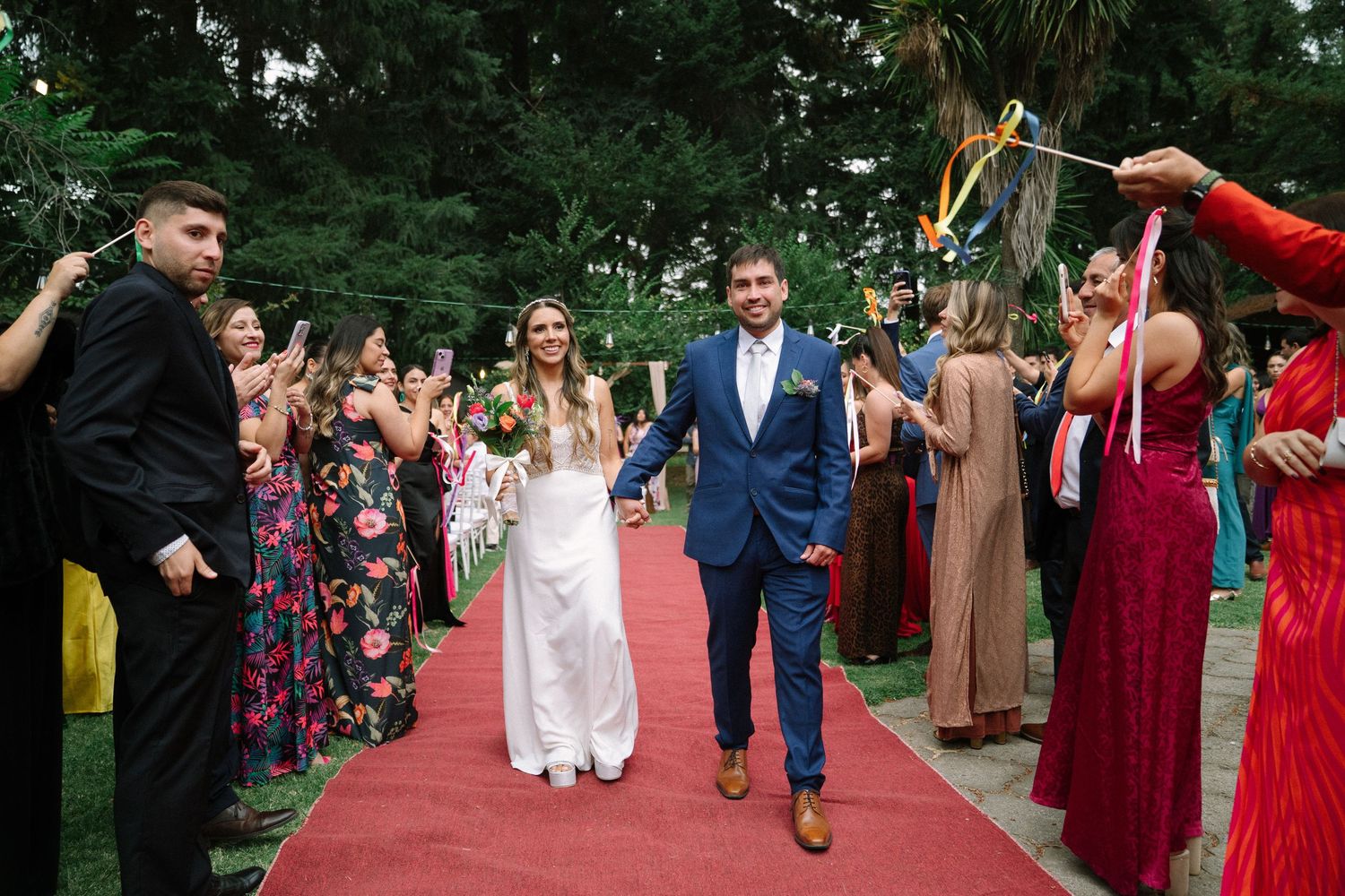 Newlyweds walk down a red carpet aisle while wedding guests celebrate with sparklers in an outdoor garden setting.