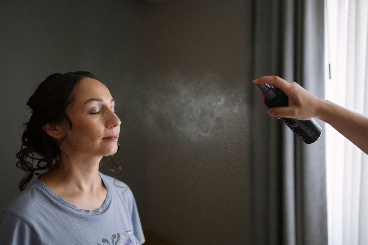 Hair stylist spraying hairspray on a bride's updo during wedding preparations in a softly lit room.