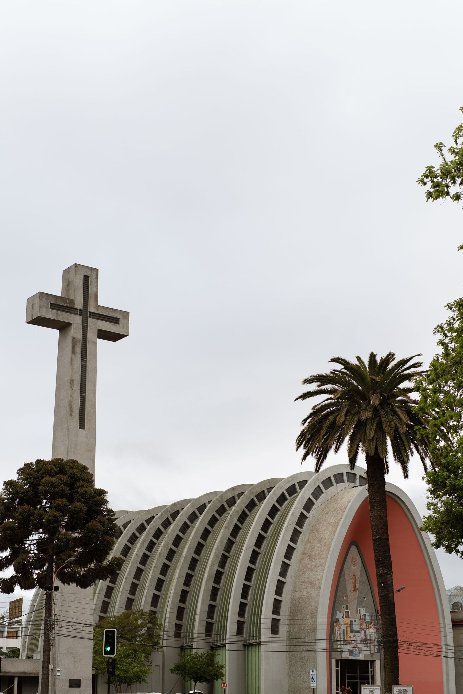 A modern church building with a large white cross and palm tree against an overcast sky.