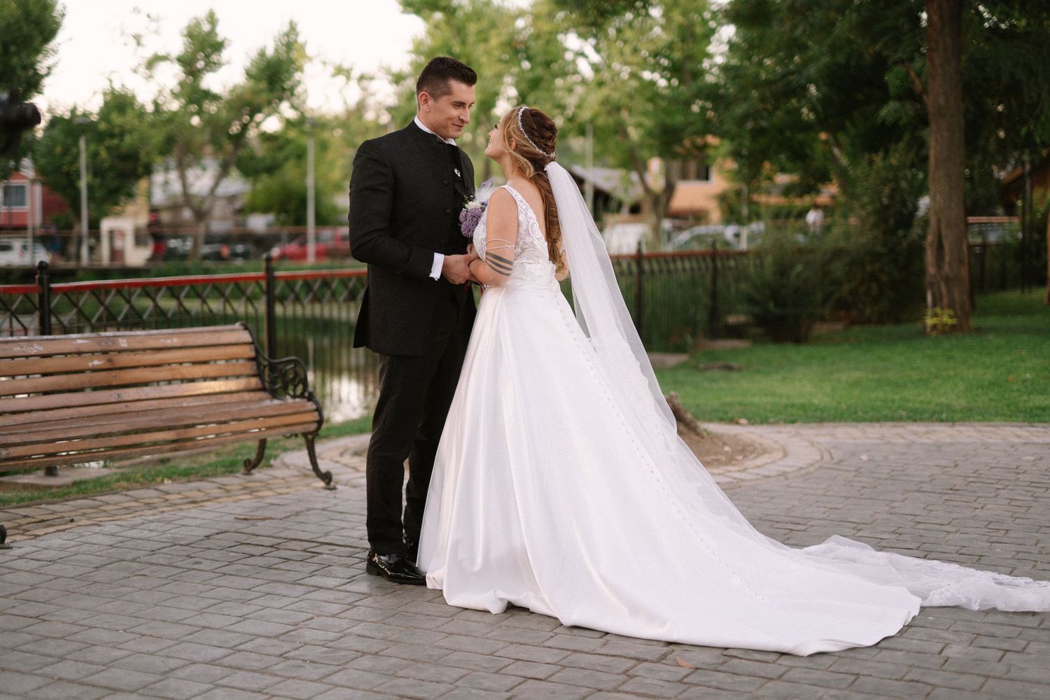 Newlywed couple in formal attire embracing in a park with trees and benches.