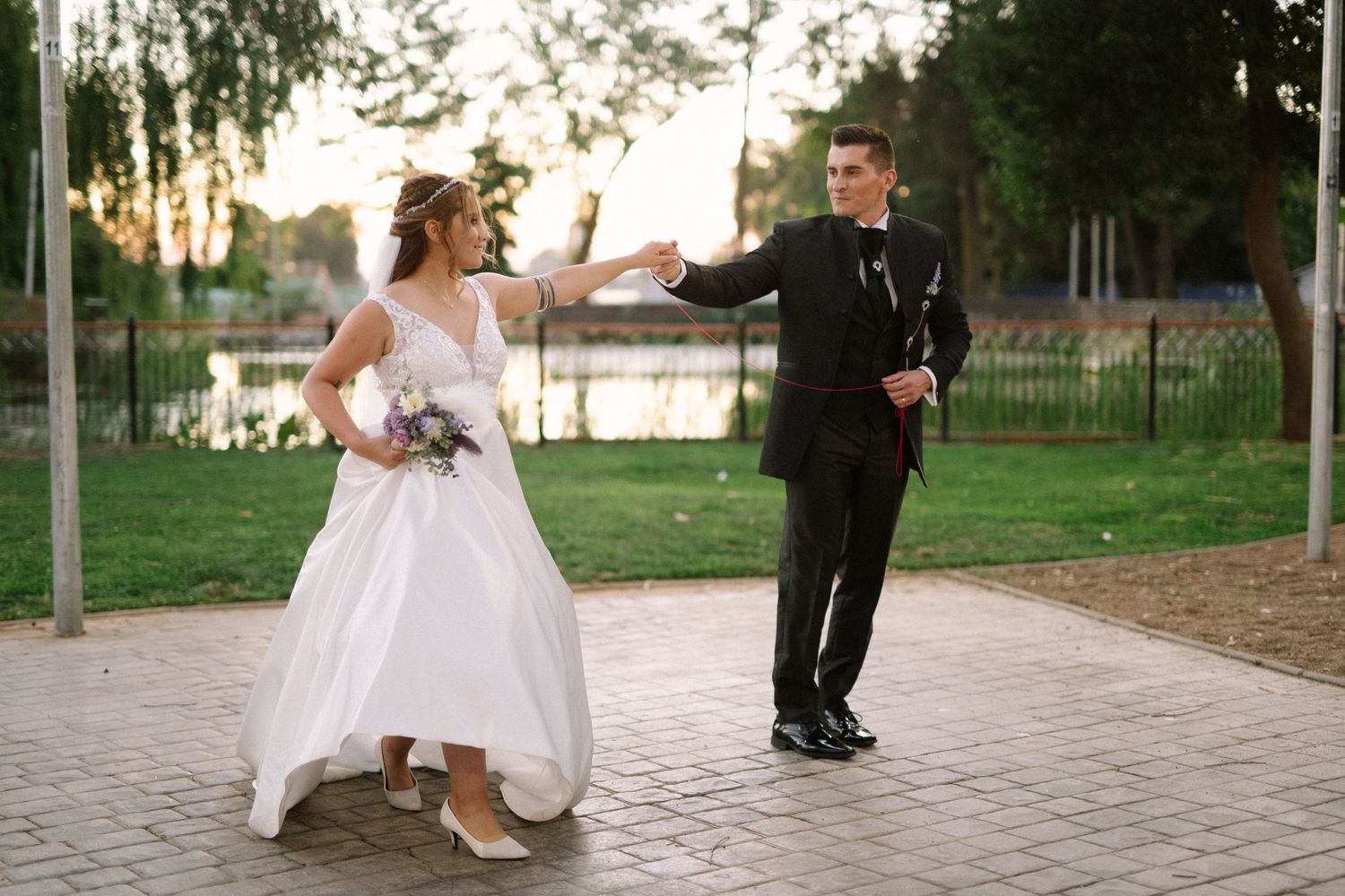 Bride and groom share a joyful dance moment on a brick pathway near a lake.