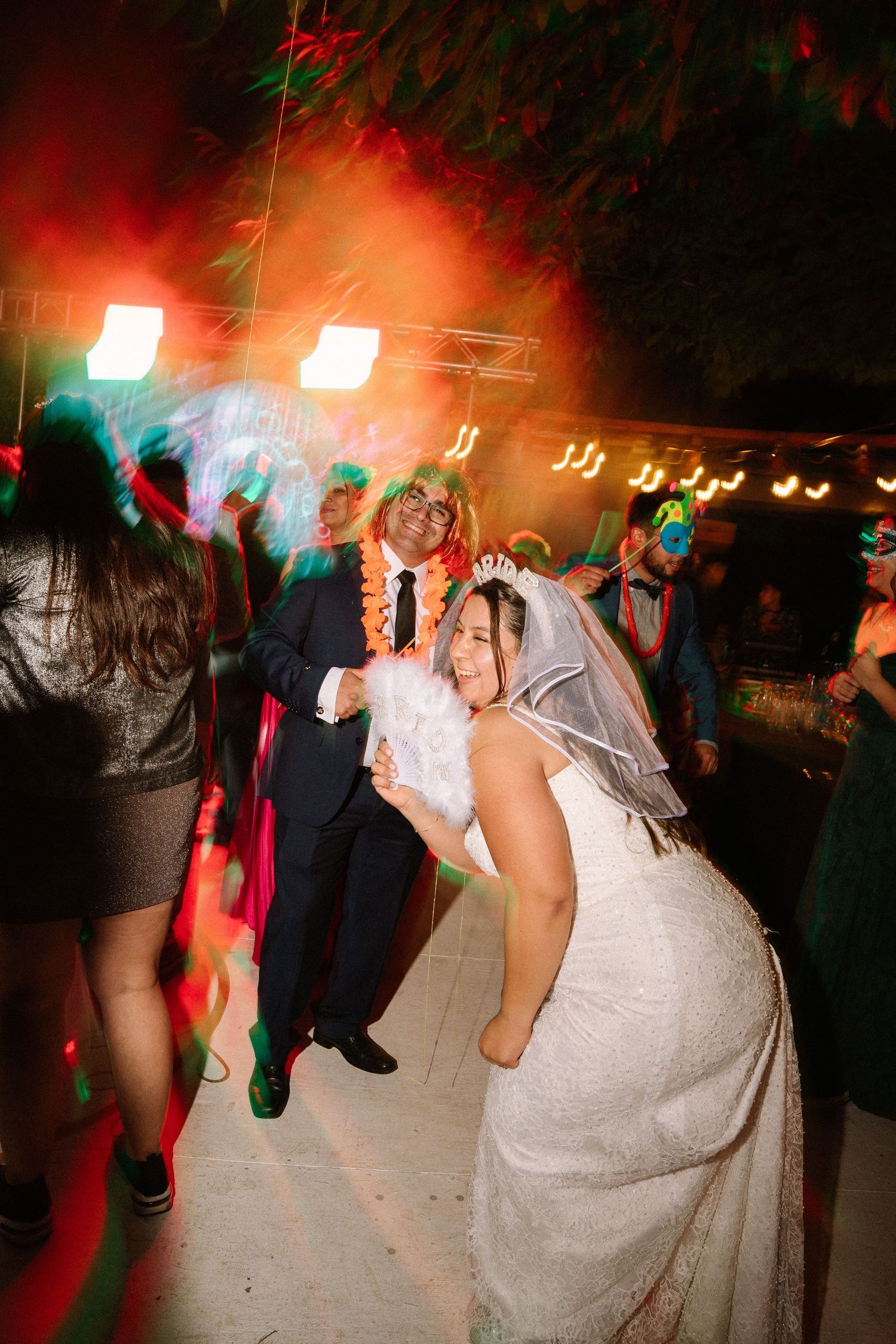 Bride in white gown and veil dances energetically with guests under colorful lights at wedding reception.