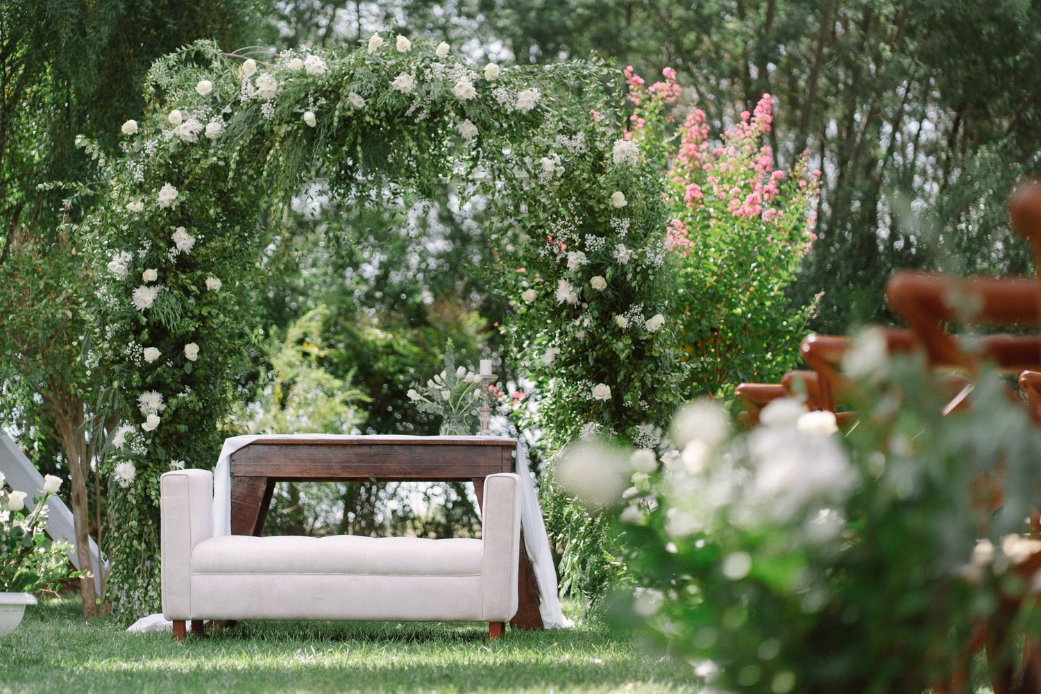 White floral arch frames a cream loveseat on lush green grass at an elegant outdoor garden wedding venue.