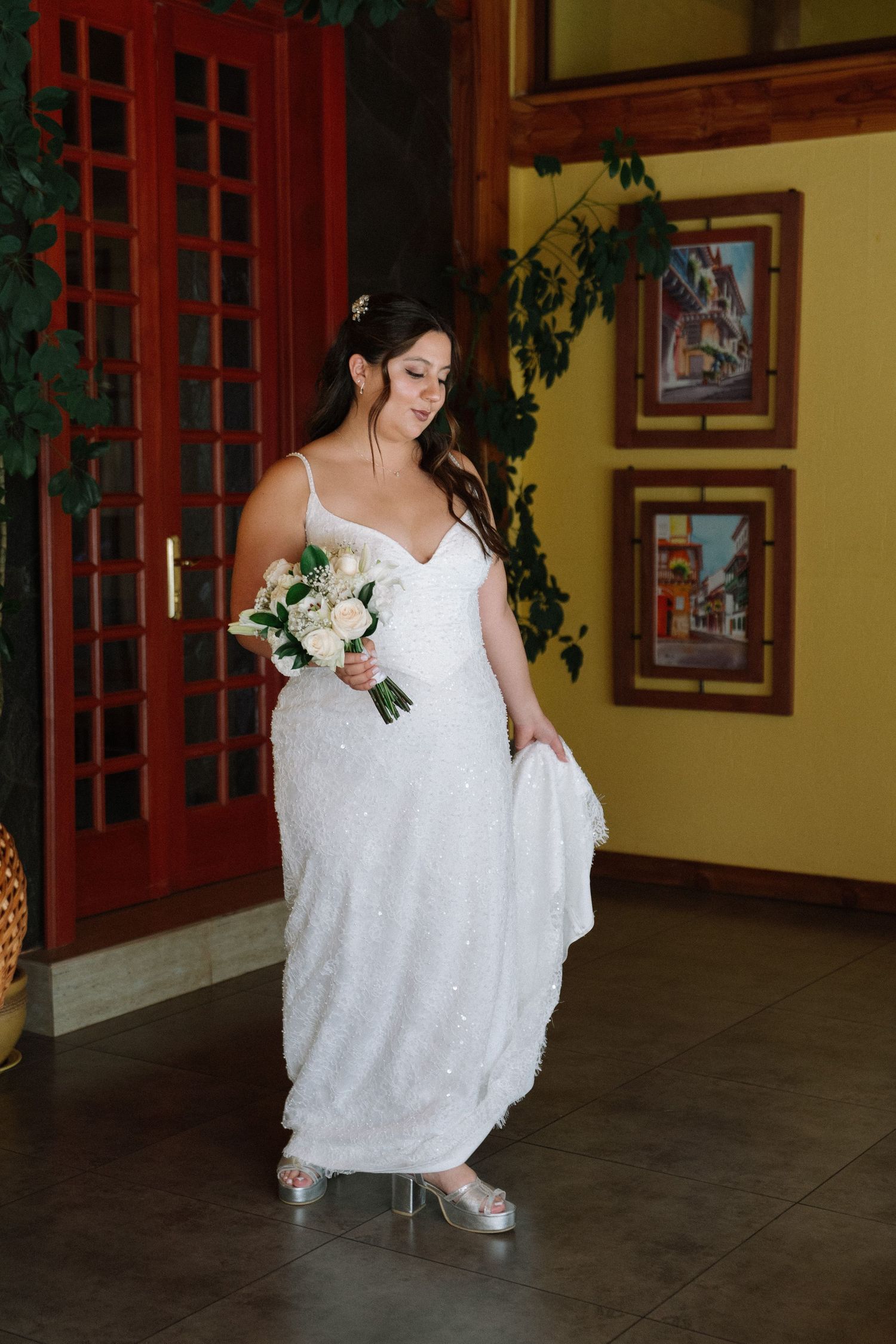 Bride in a beaded white gown holding a white bouquet stands in a warmly decorated venue interior.