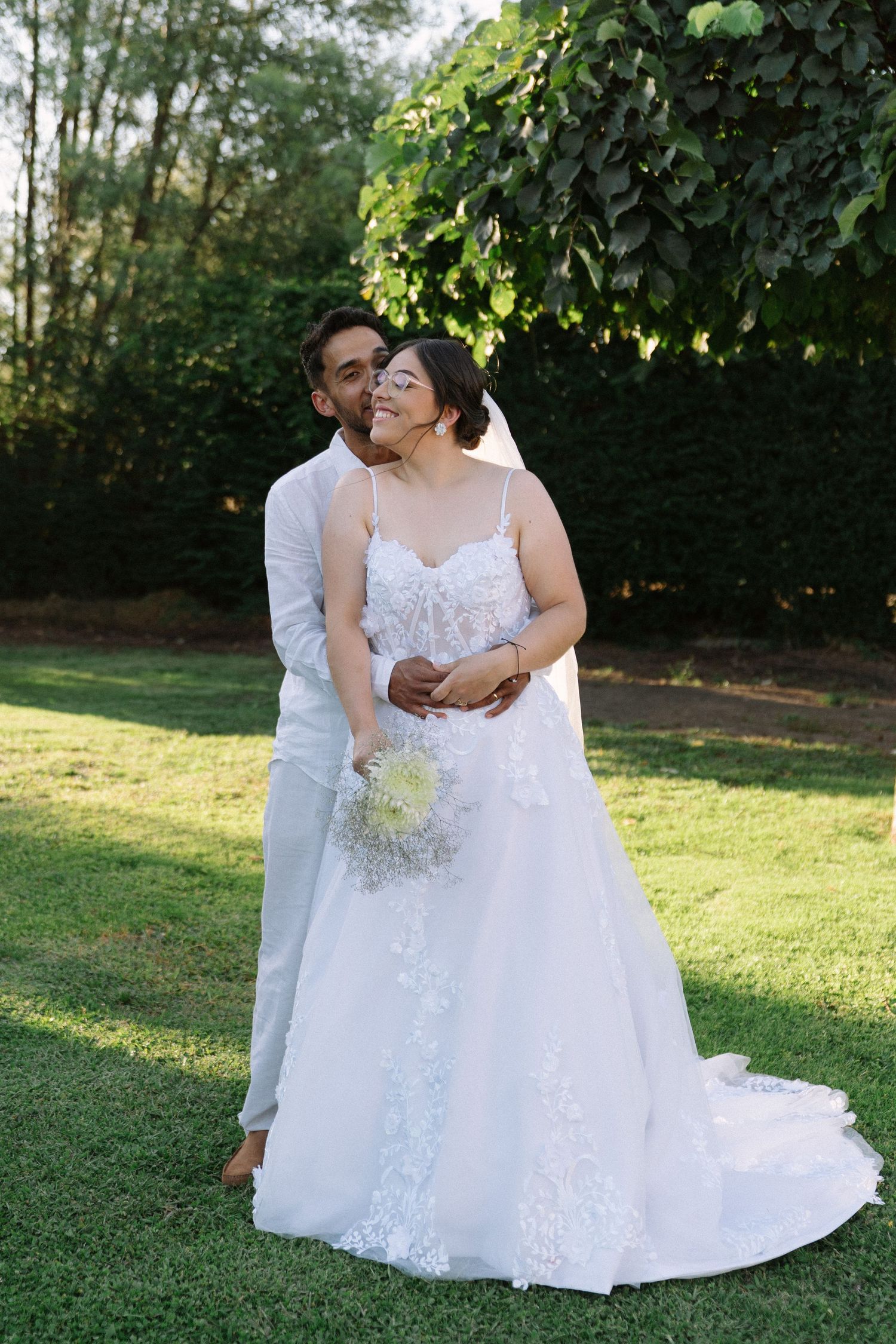 Groom kisses bride on cheek as she holds white bouquet in sunny garden.