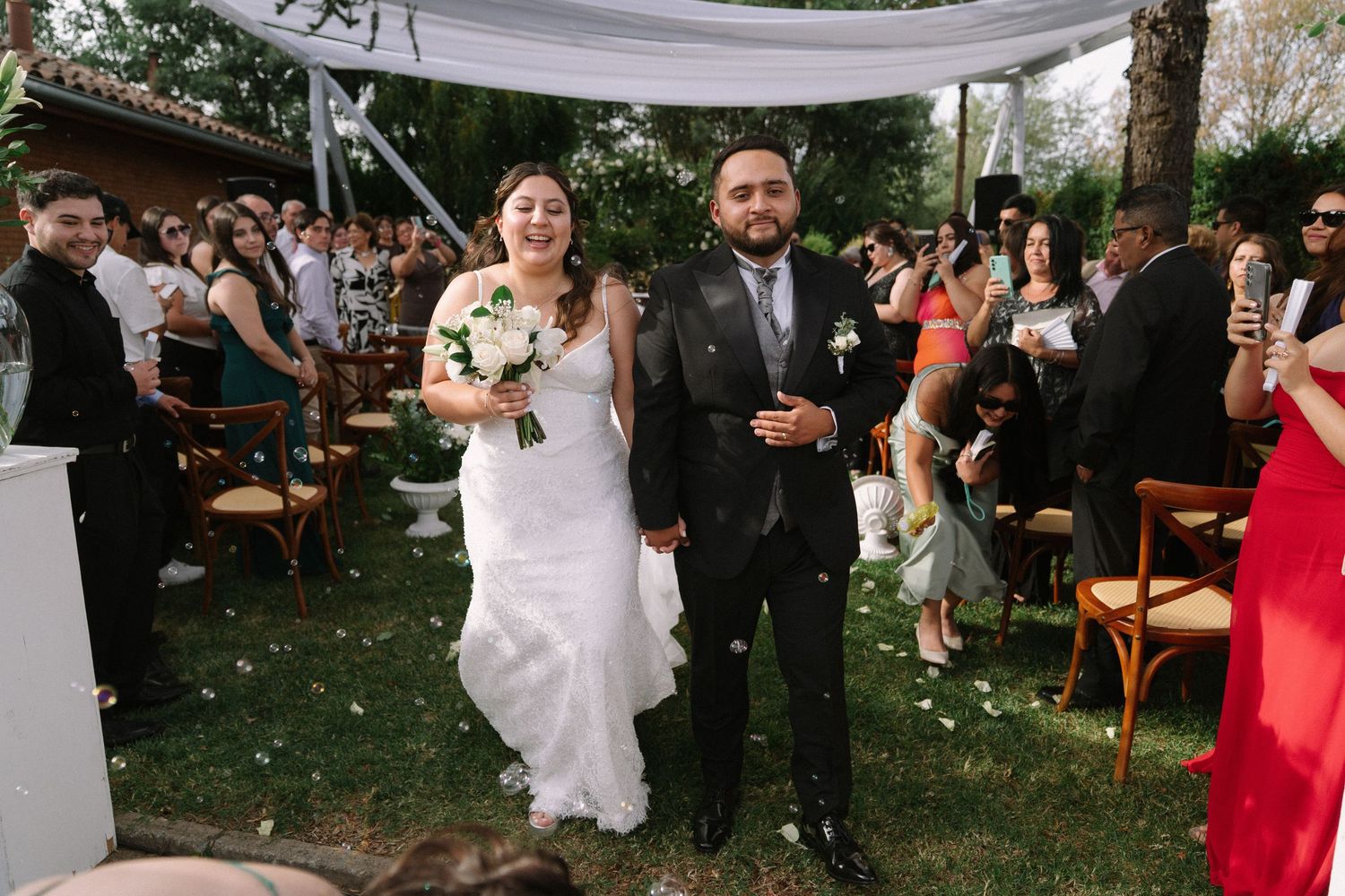 Happy newlywed couple walking down outdoor wedding aisle surrounded by cheering guests in garden.