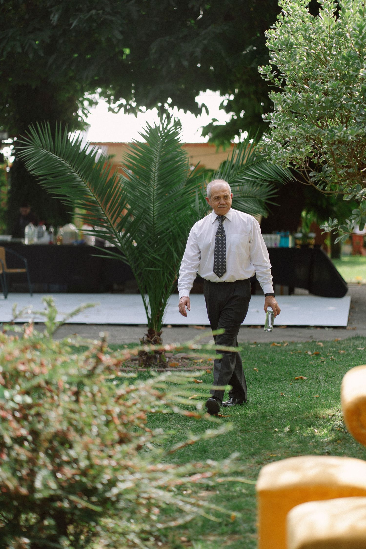 Elderly man in white shirt and tie walking through lush green garden at outdoor wedding.