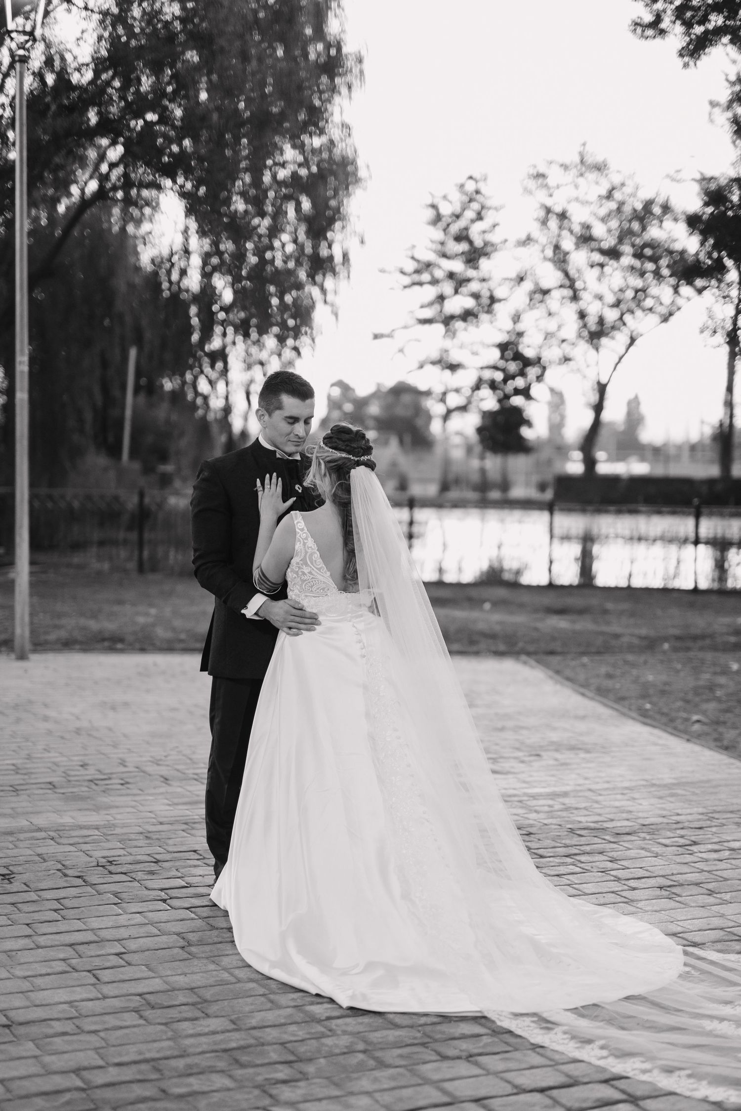 A black and white photo of a bride and groom embracing on a brick path beside a lake at sunset.