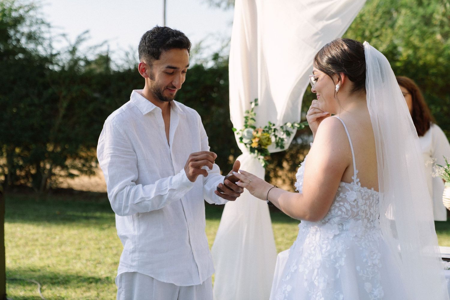 Groom in white shirt placing wedding ring on brides finger during outdoor ceremony with floral arch backdrop.