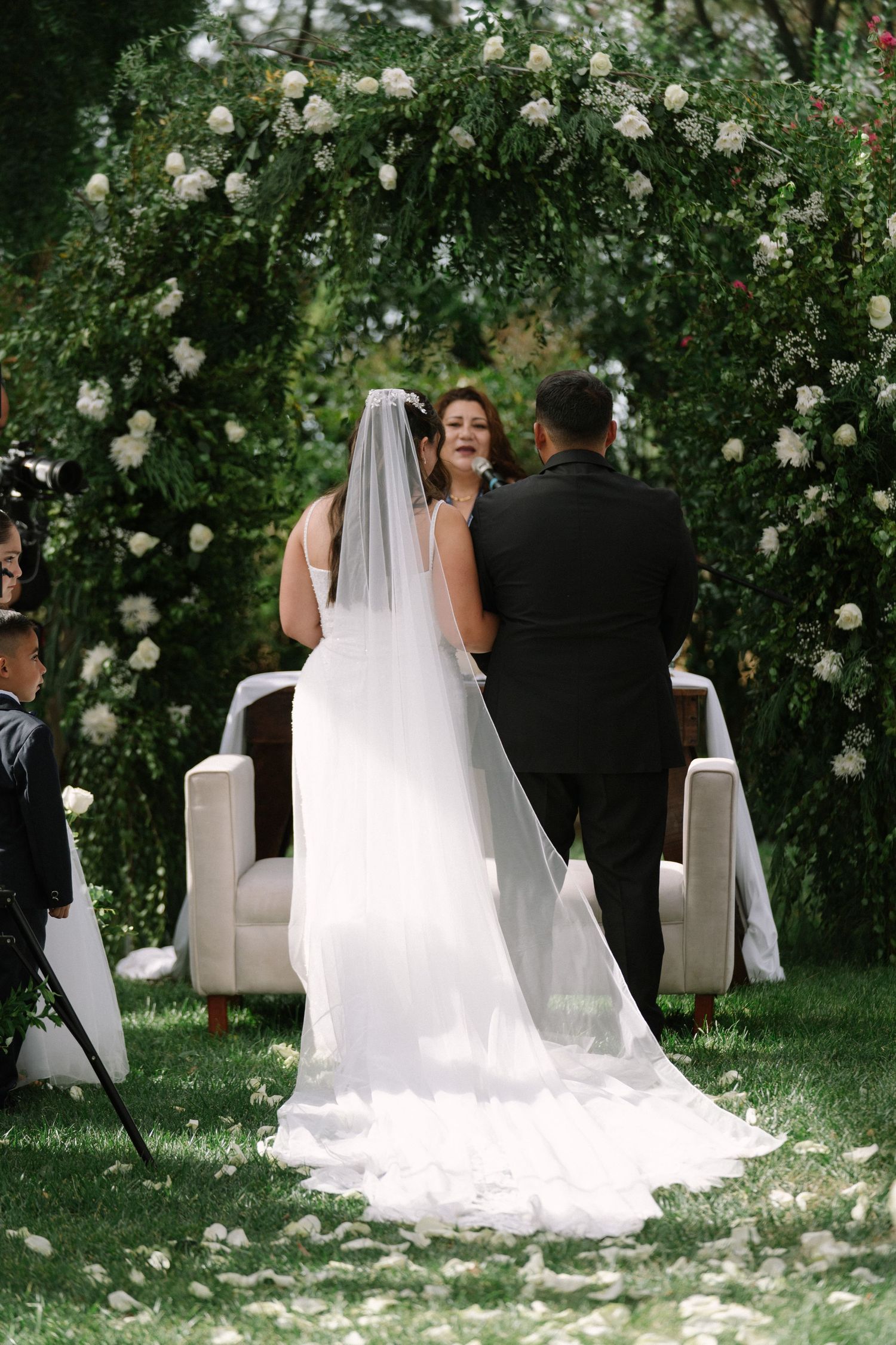 Bride and groom exchanging vows under a lush green floral arch at an outdoor ceremony.