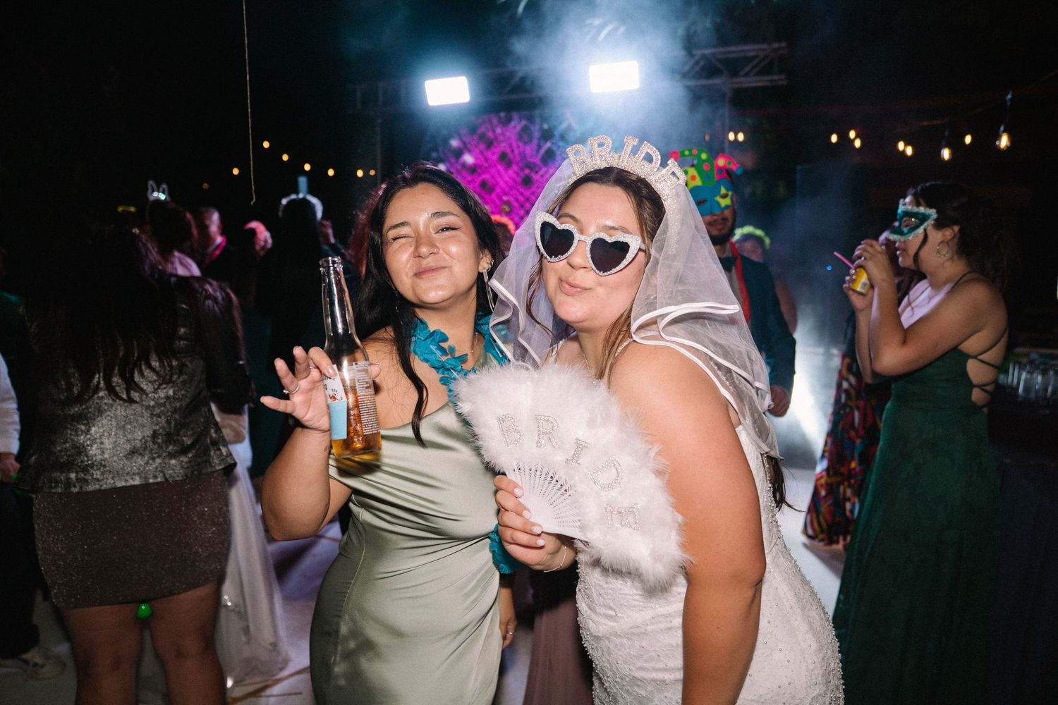 Two women pose playfully at wedding reception, one in bridal veil and fur wrap with heart sunglasses.