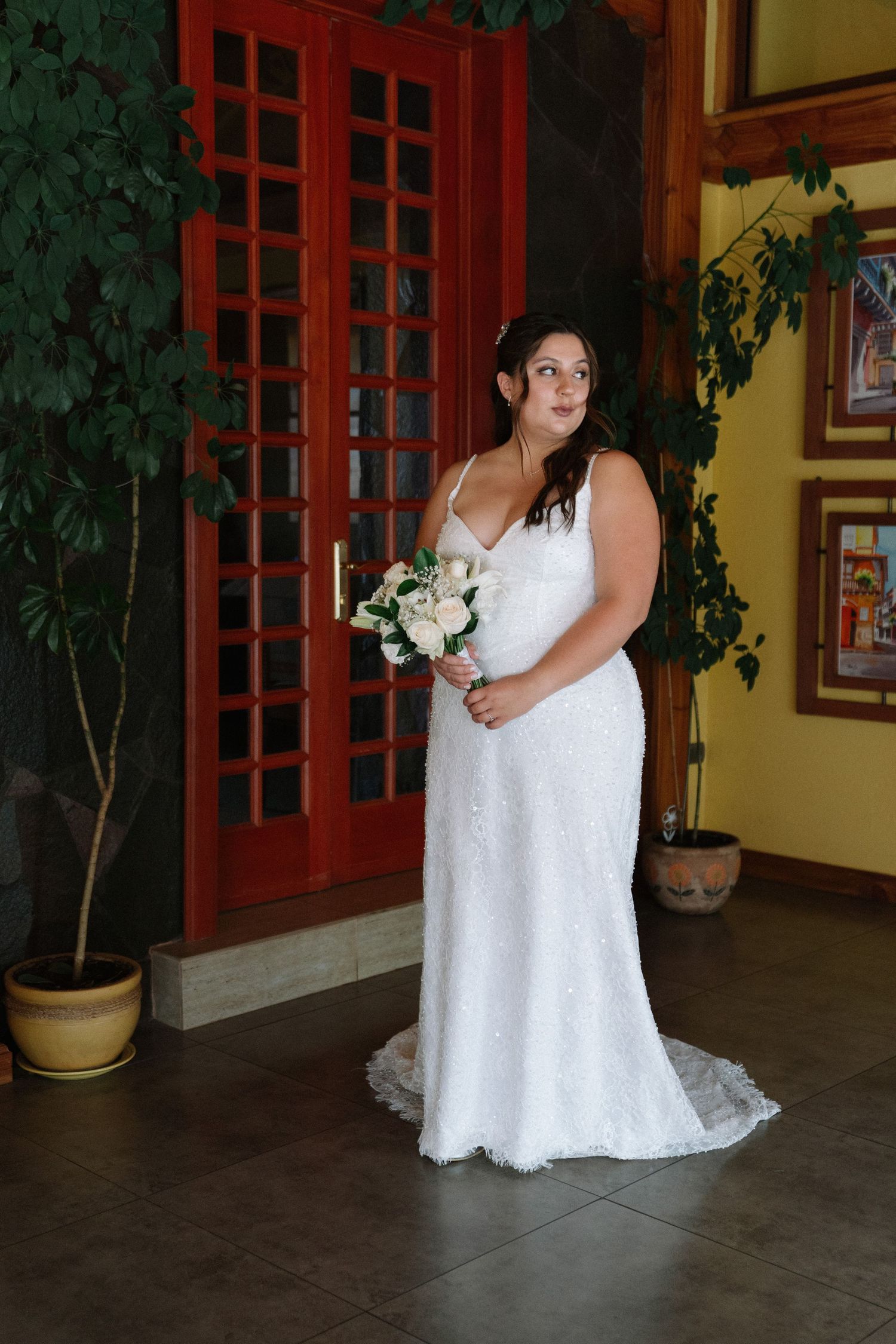 Bride in a white lace sequined gown holding a bouquet of pink and white roses indoors.