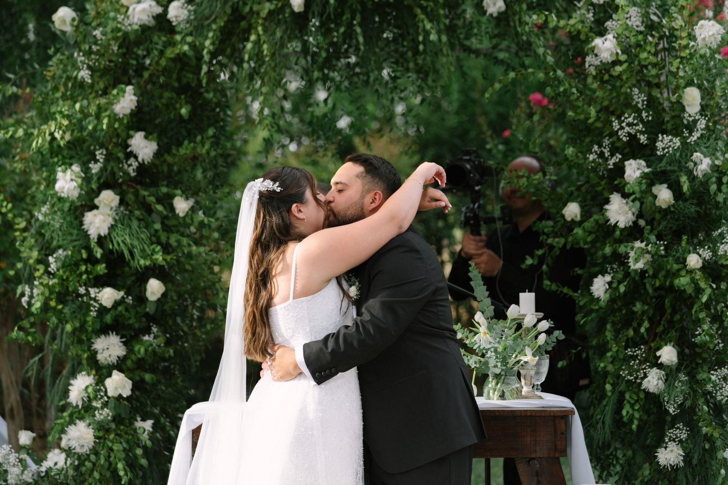 Bride and groom share their first kiss beneath a lush green floral arch at their outdoor wedding ceremony.