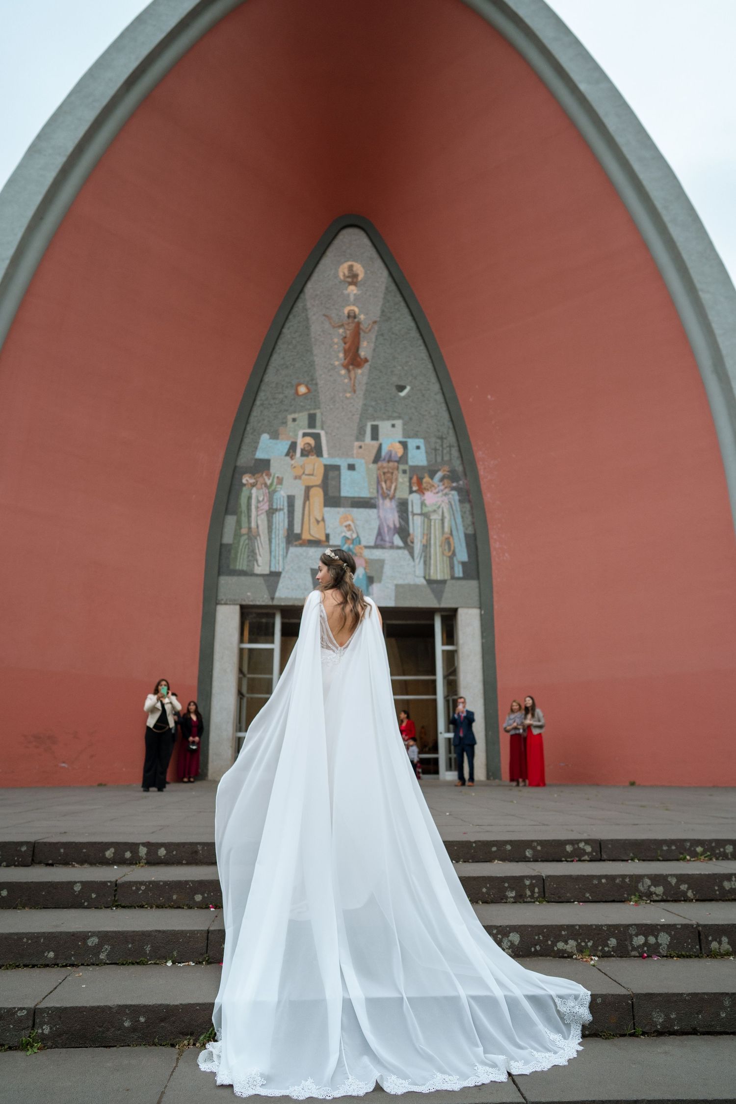 A bride in a flowing cape-like veil stands on steps before a red-arched church entrance.
