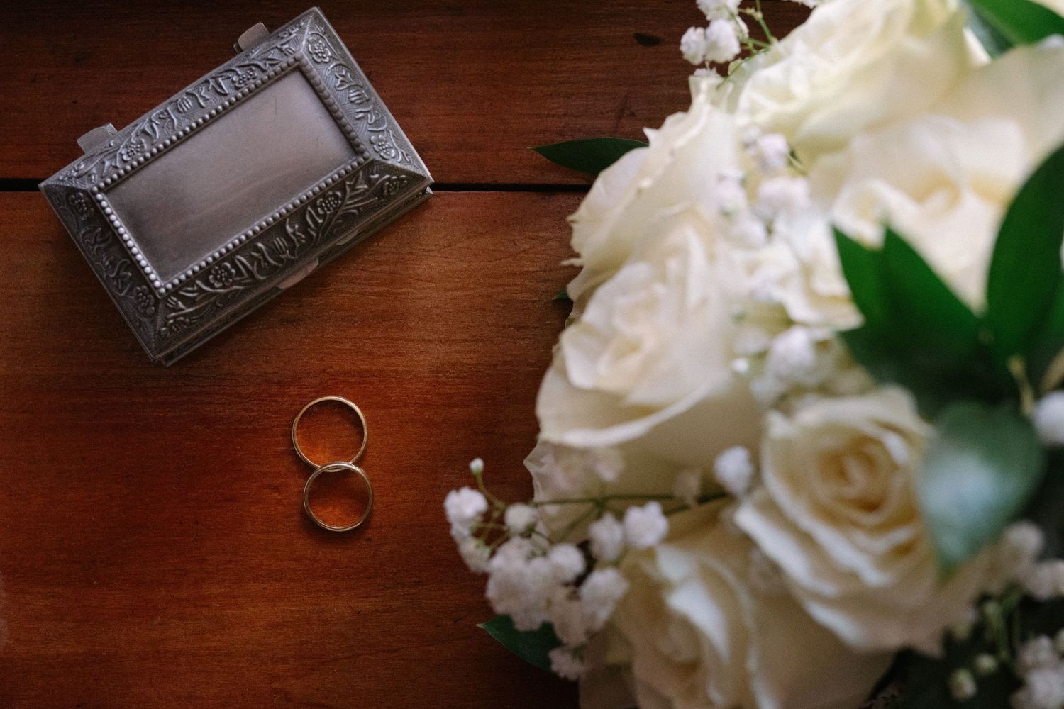 Wedding rings and ornate frame displayed with white rose bouquet on wooden surface.