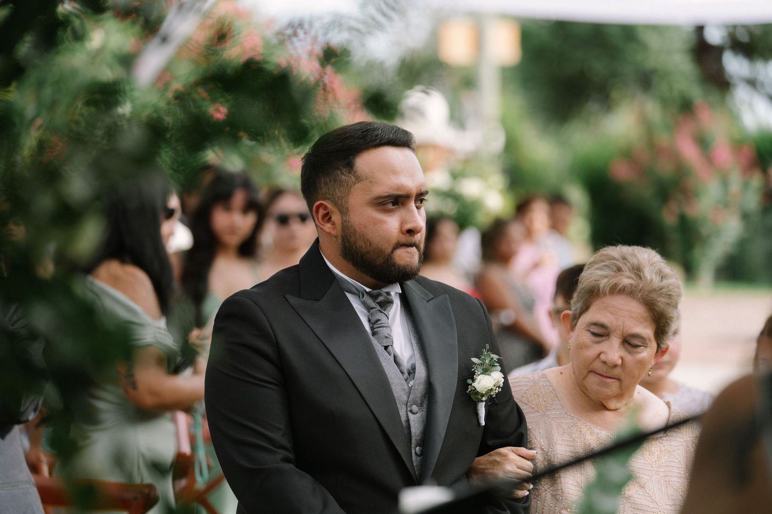 Groom in black suit with boutonniere standing at outdoor wedding ceremony aisle with guests.