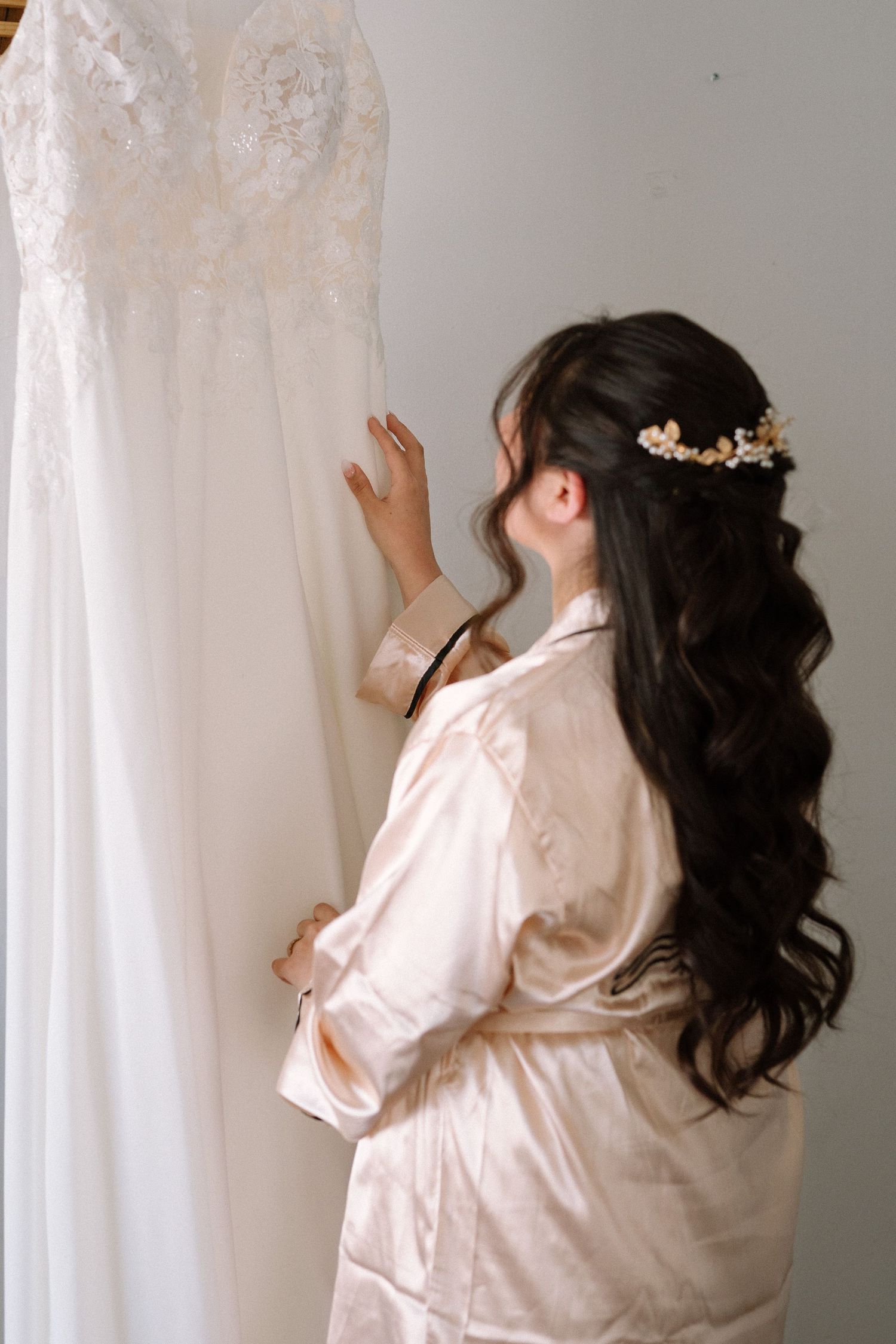Figure in silk robe admiring white lace wedding dress hanging on wall.