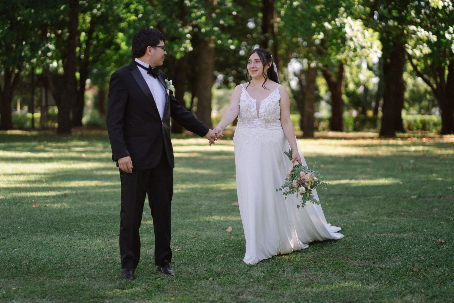 A wedding couple shares a playful moment on a sunlit lawn surrounded by trees.