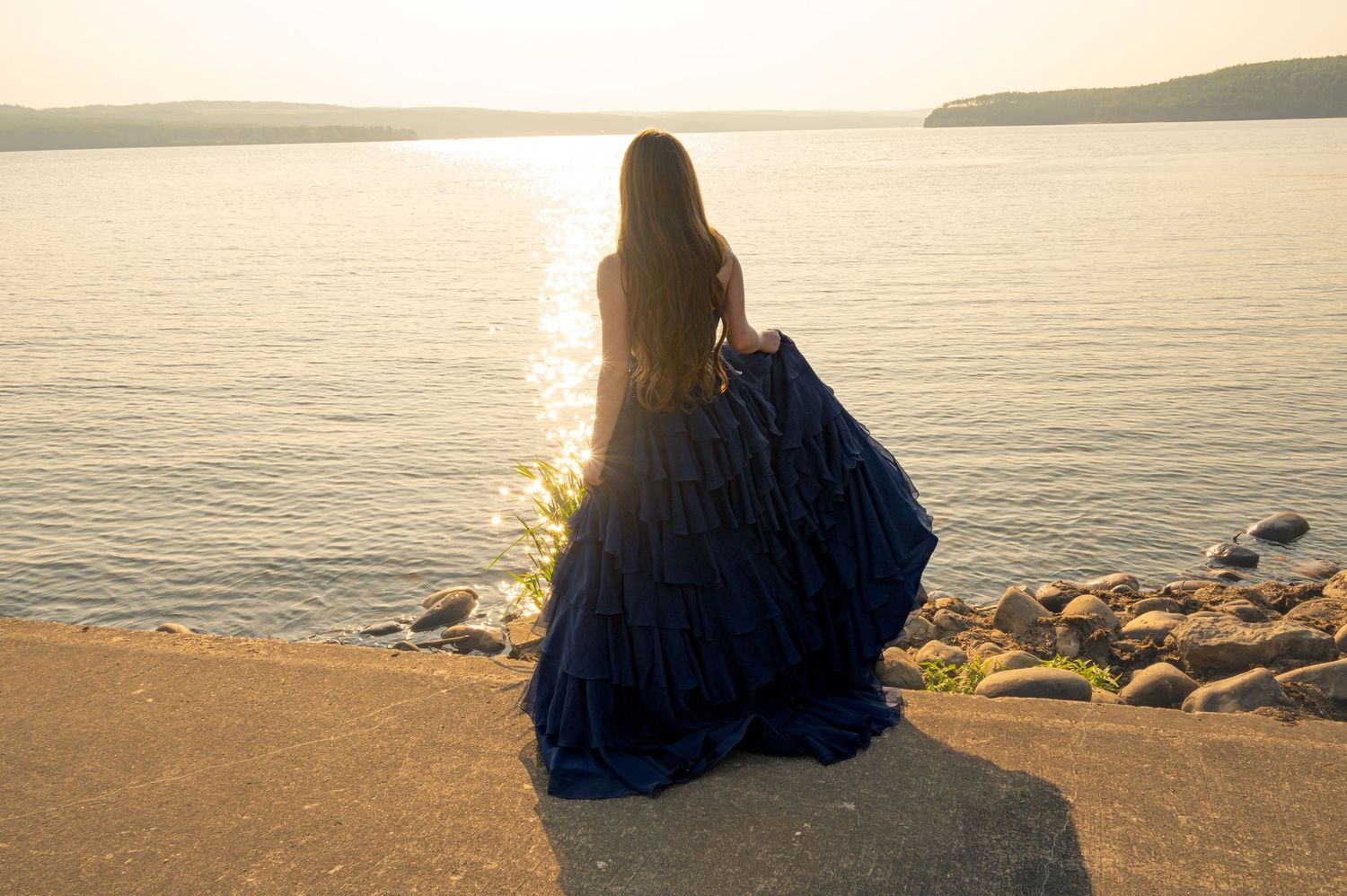 A figure in a flowing navy blue gown stands on a rocky beach at sunset overlooking calm waters.