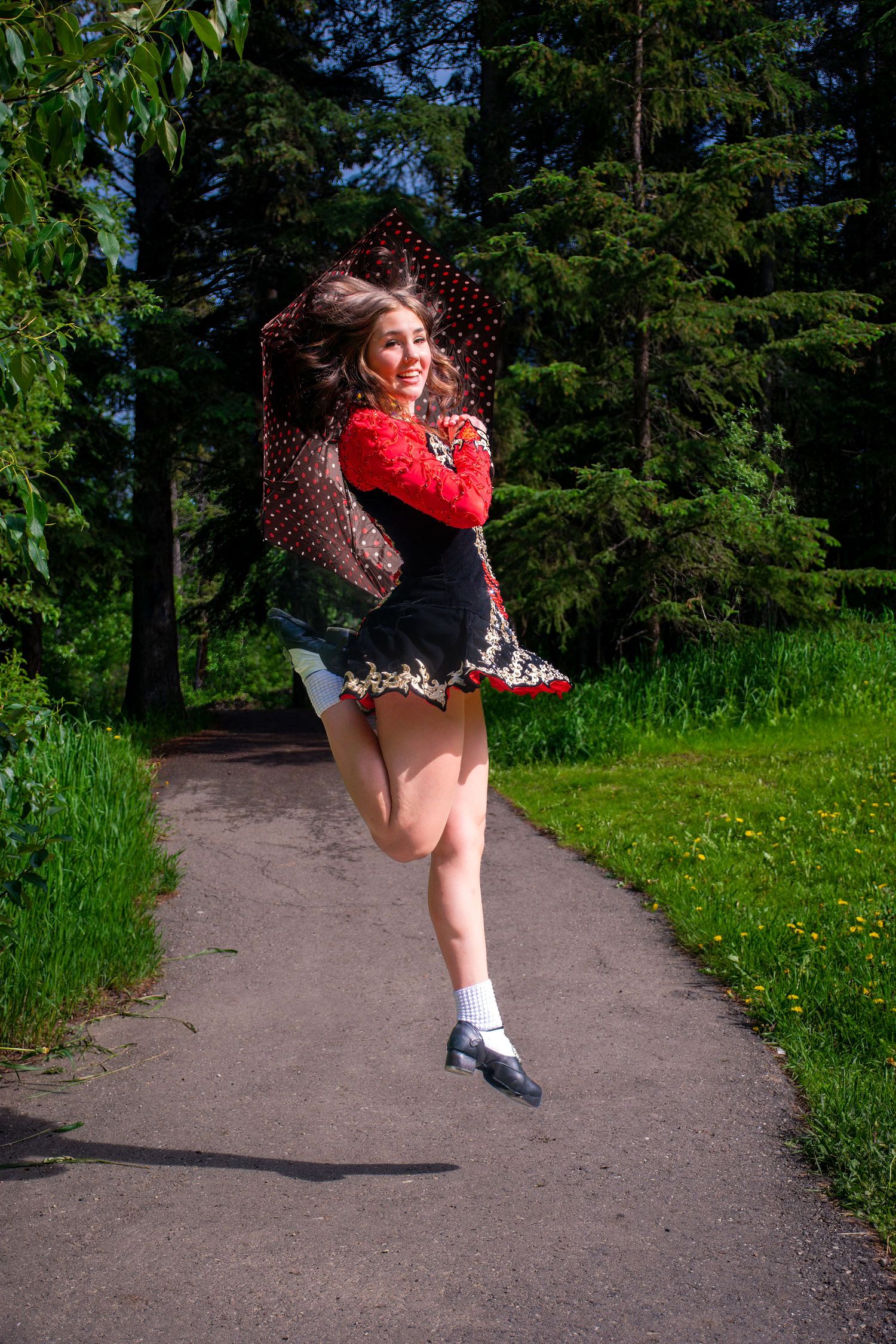 A dancer in a red top leaps gracefully on a park path surrounded by greenery.