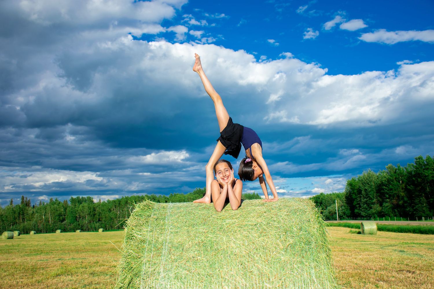 A dancer performs a backbend on a hay bale against a dramatic cloudy sky.