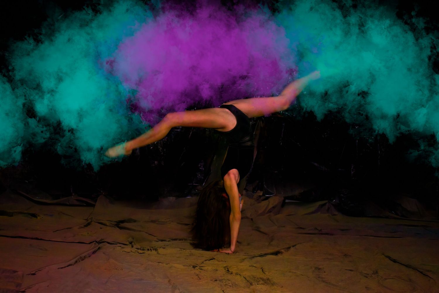 Acrobatic handstand pose silhouetted against turquoise and purple powder clouds.