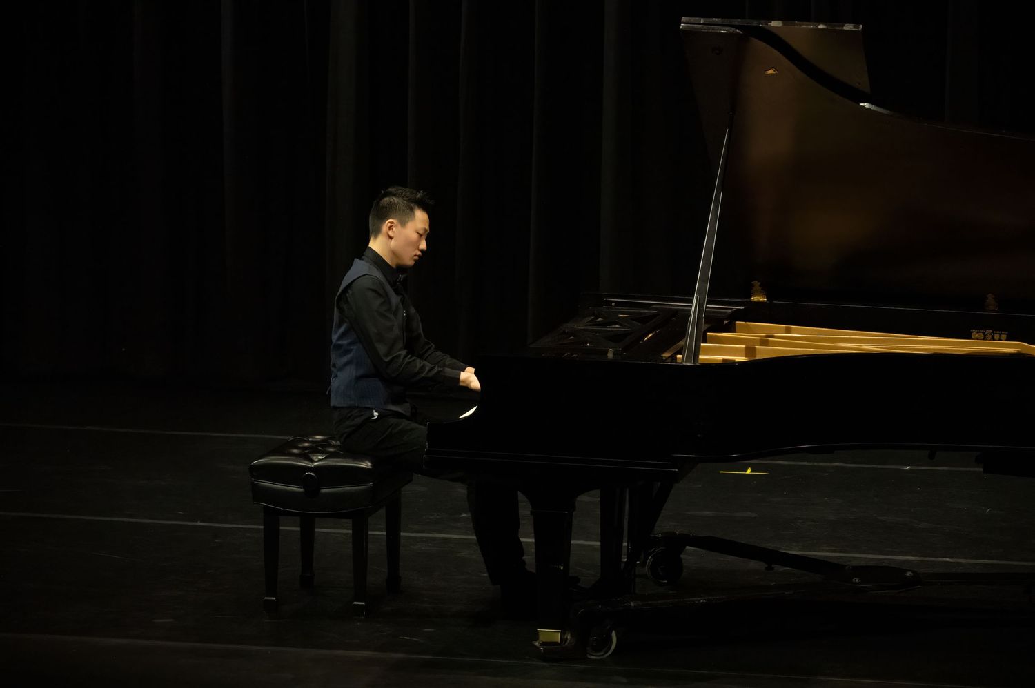 A pianist performs on a grand piano on a dark stage illuminated by dramatic lighting.