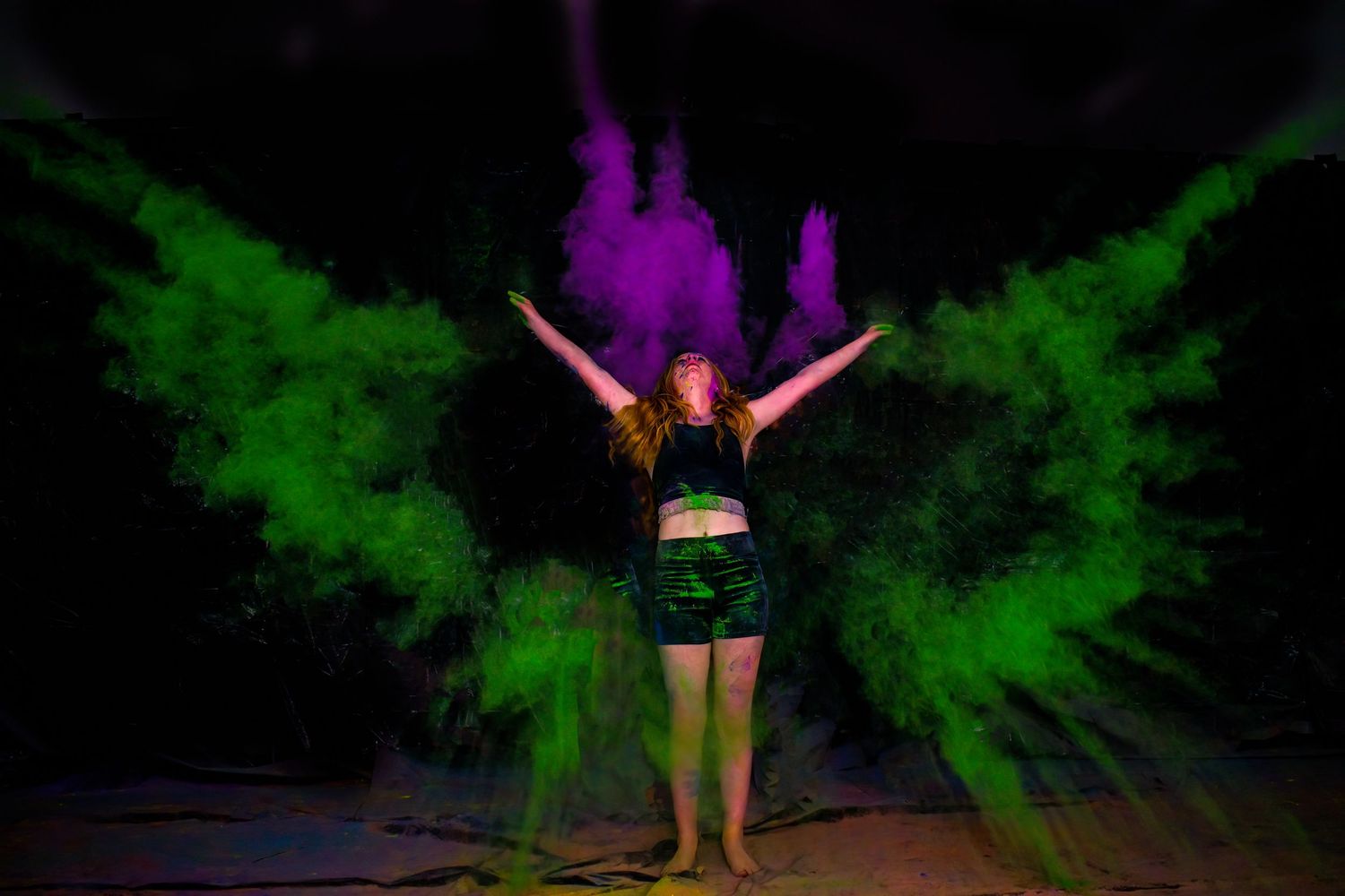 Performer surrounded by vibrant green and purple powder clouds during dynamic dance pose.