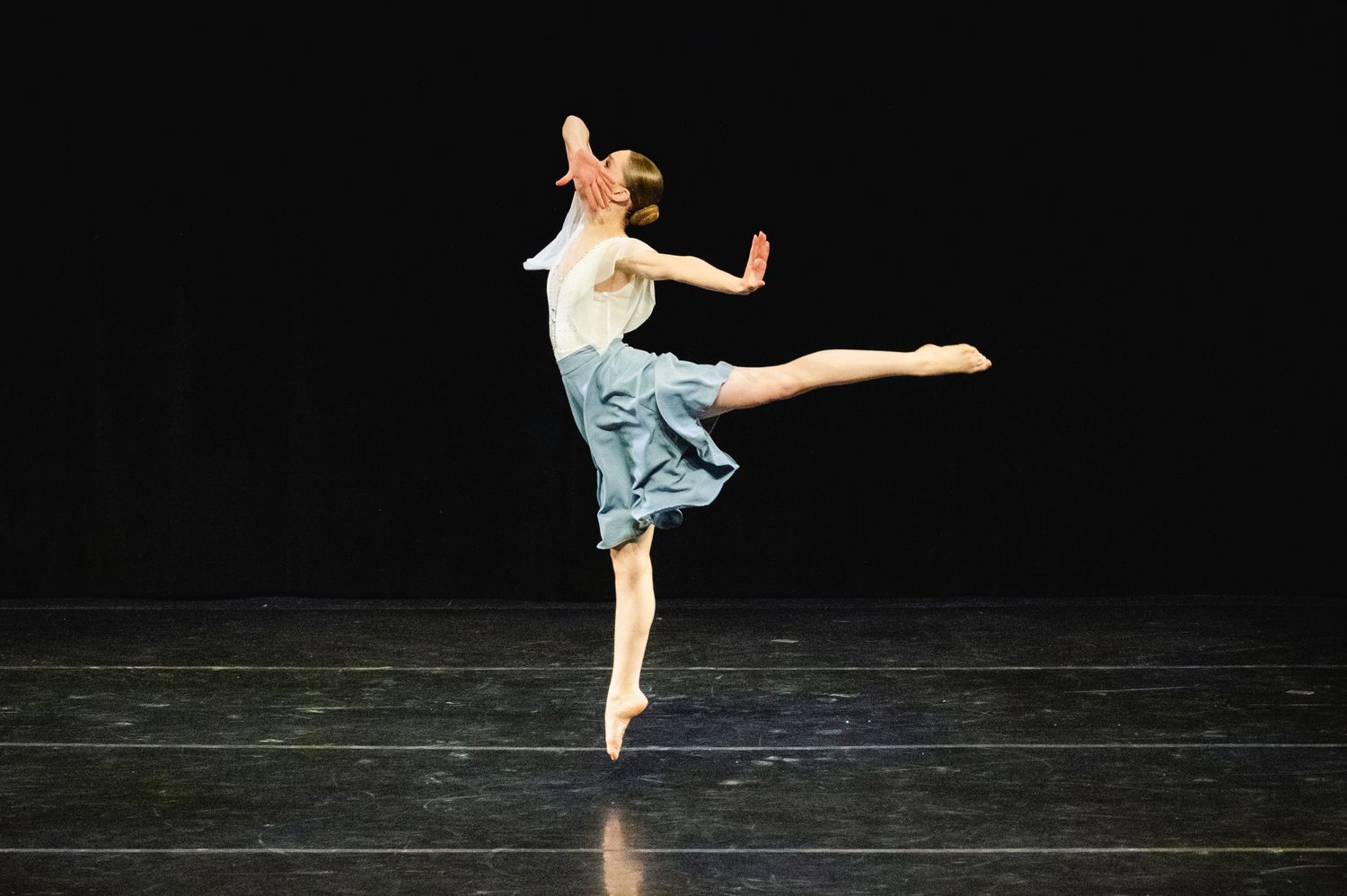 Dancer in blue denim skirt and white top performs an artistic dance move on dark stage.