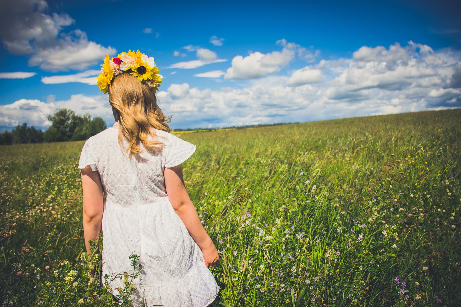 A figure in a white dress and sunflower crown stands in a sunny meadow with blue skies and puffy clouds overhead.
