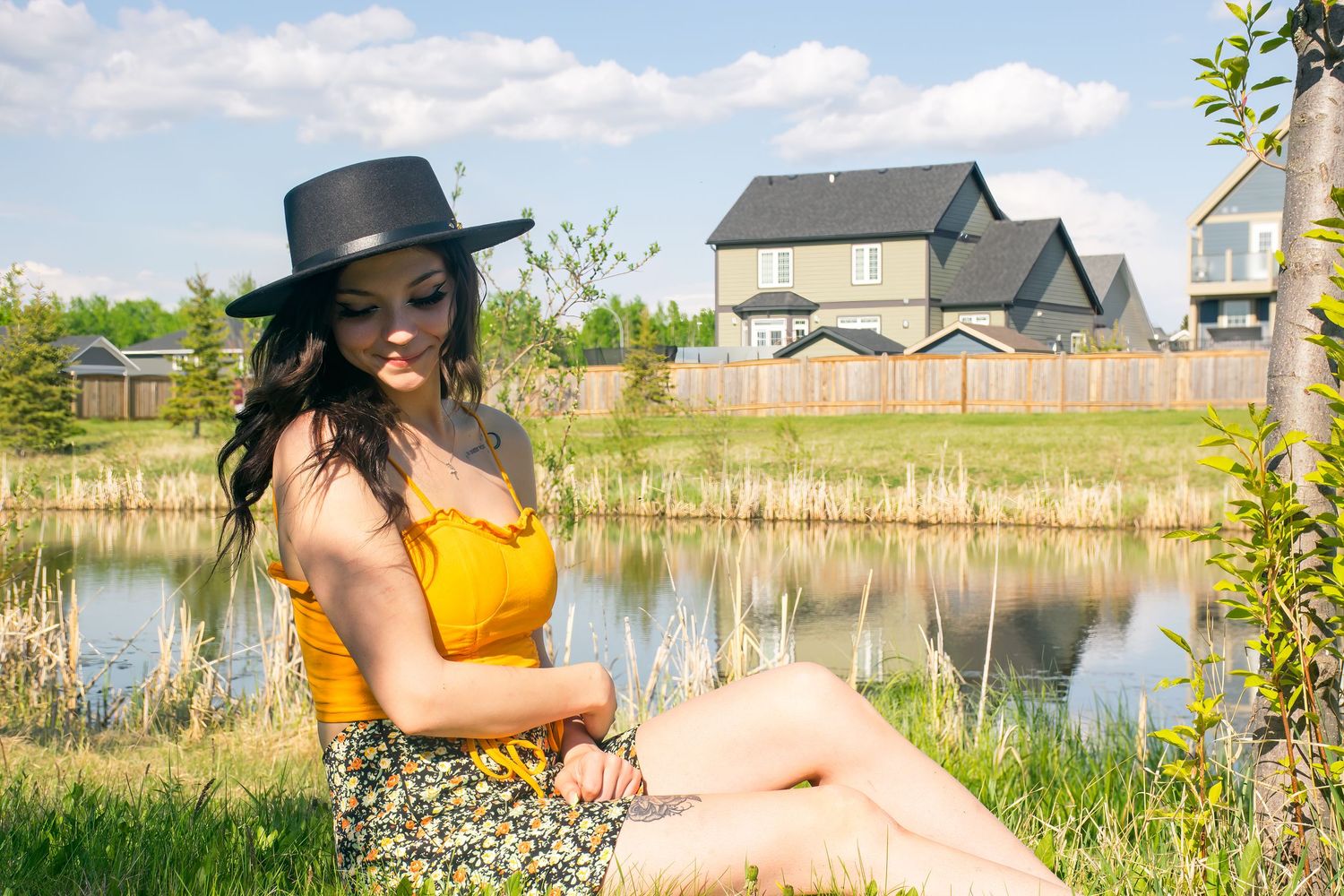 Woman in yellow top and hat poses by a pond with houses in background.