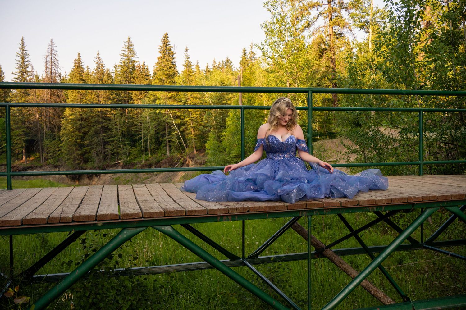 A blue gown billows elegantly across a wooden deck surrounded by tall evergreen trees at golden hour.