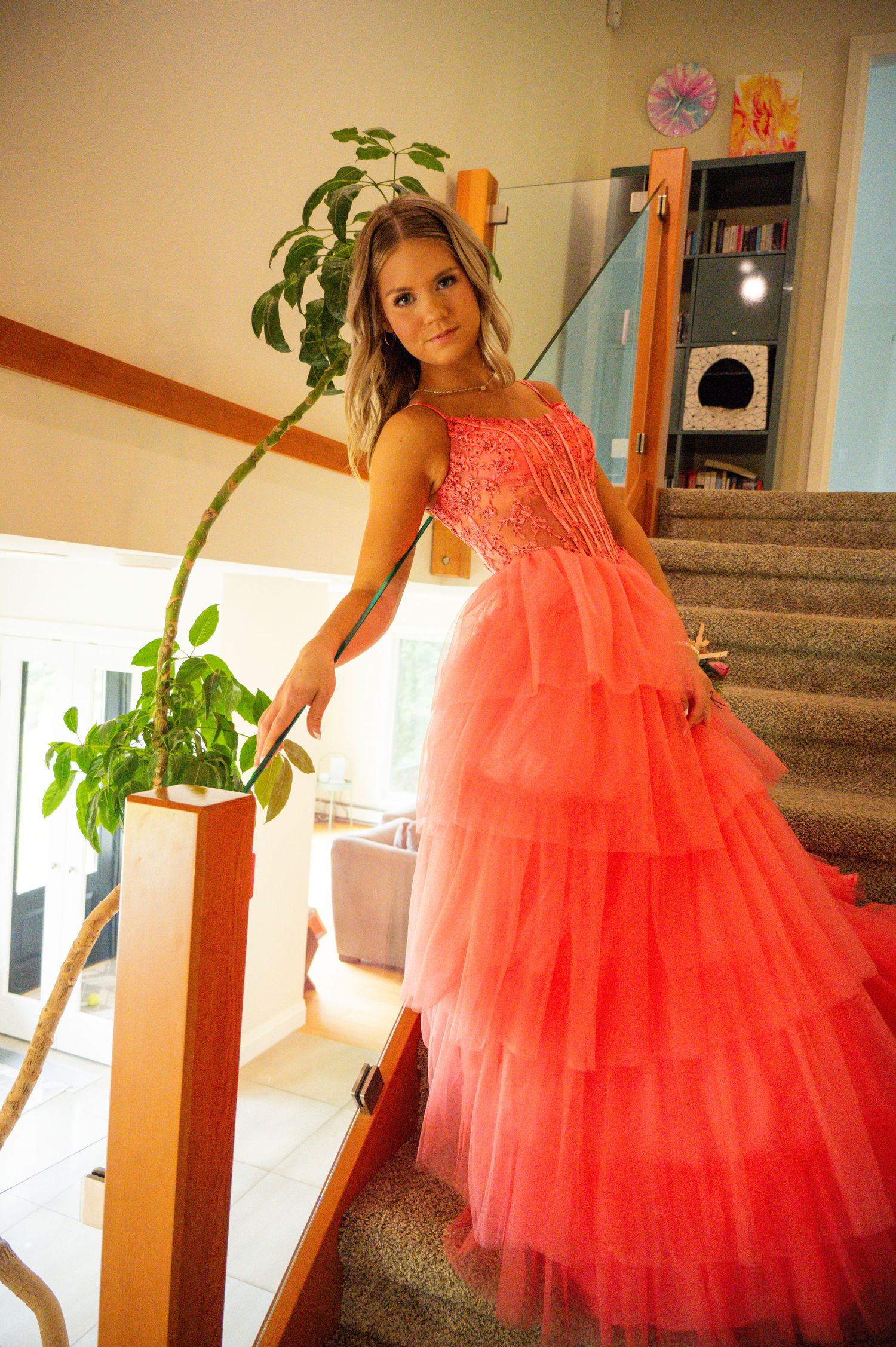 A flowing coral ballgown with tiered tulle skirt photographed on a staircase next to a potted plant.