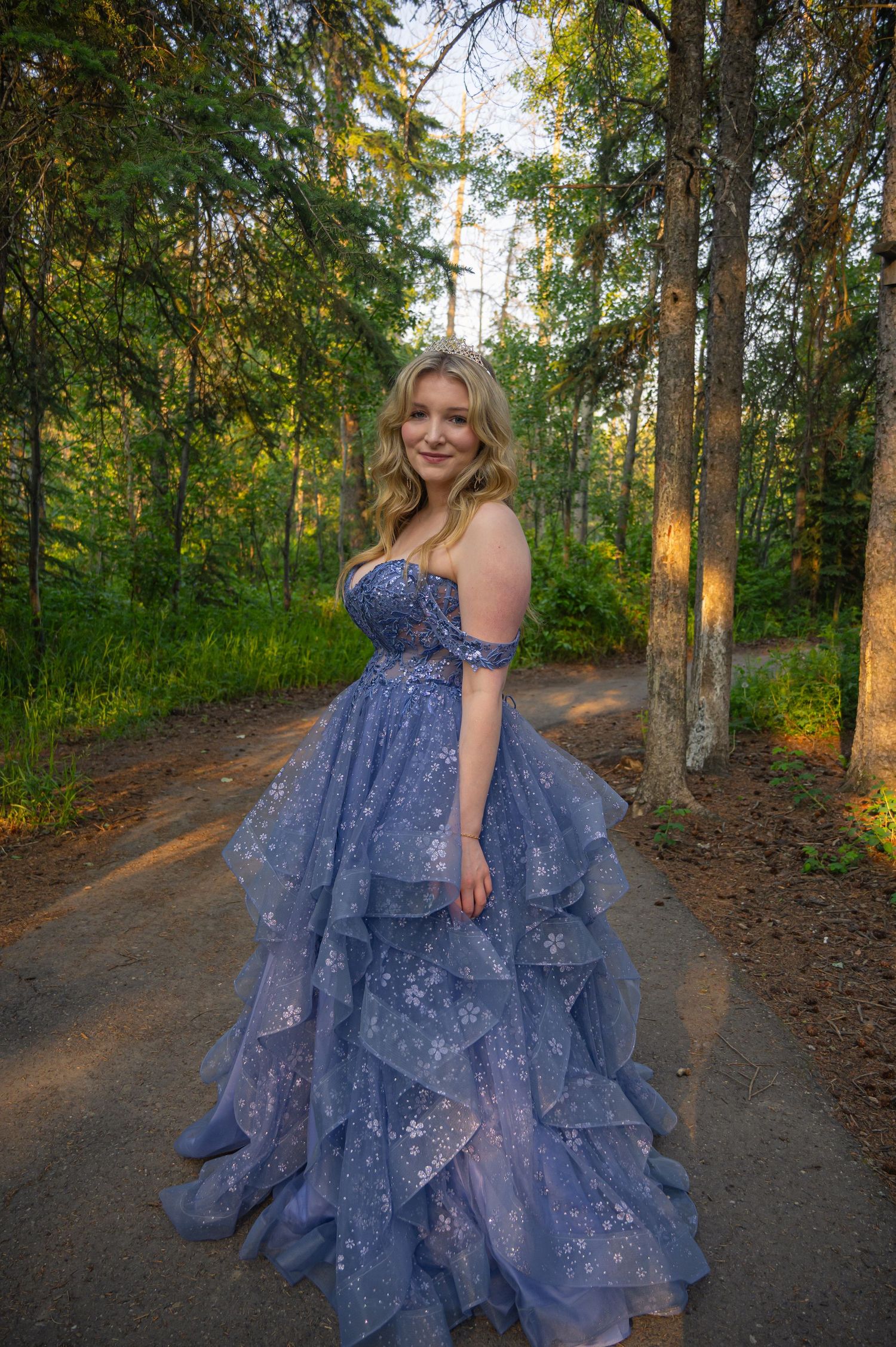 A person in a sparkling blue evening gown poses on a forest path illuminated by golden sunlight.