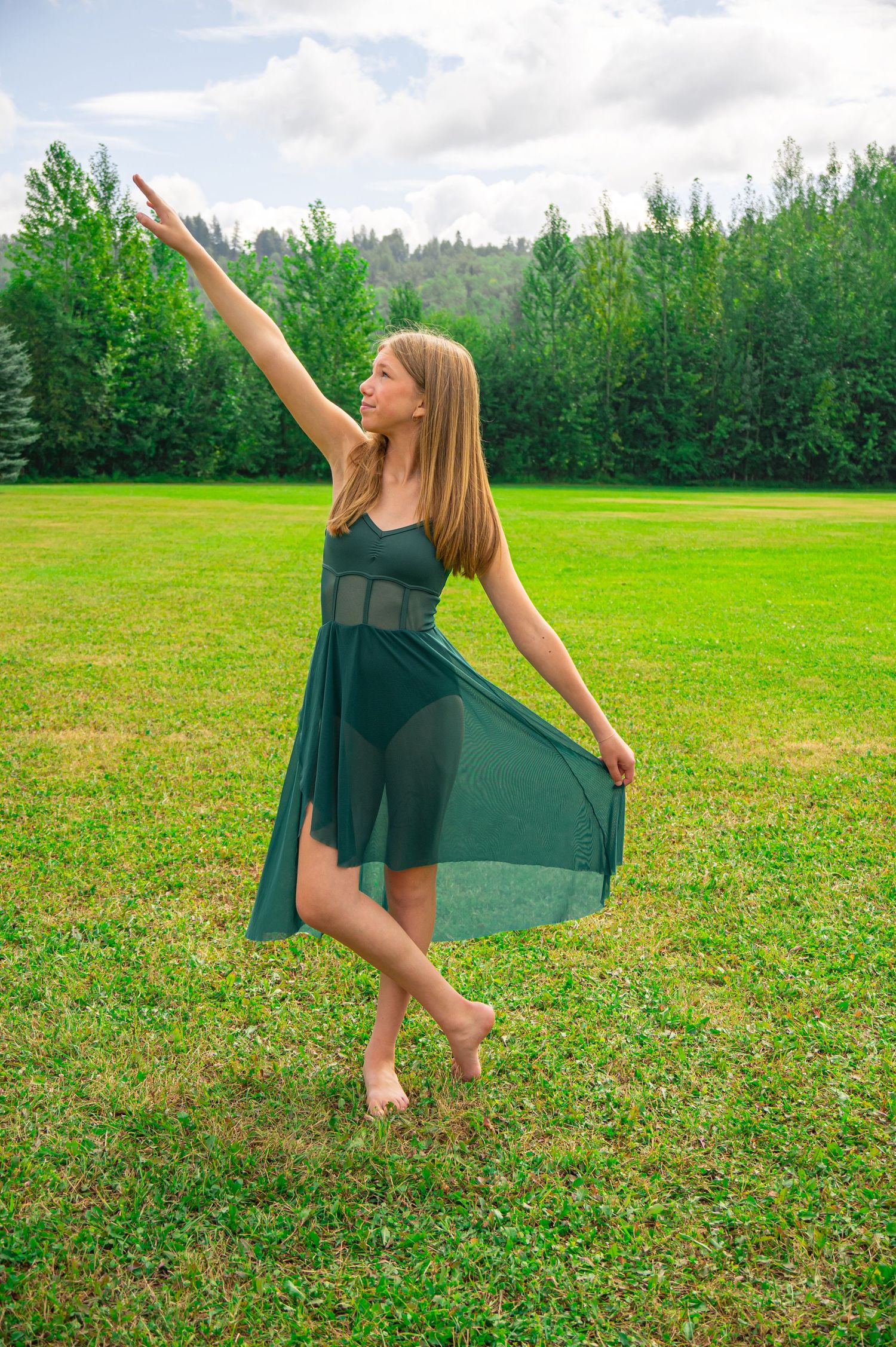 Dance performer in flowing teal dress strikes an elegant pose with raised arm on green lawn with forest backdrop.