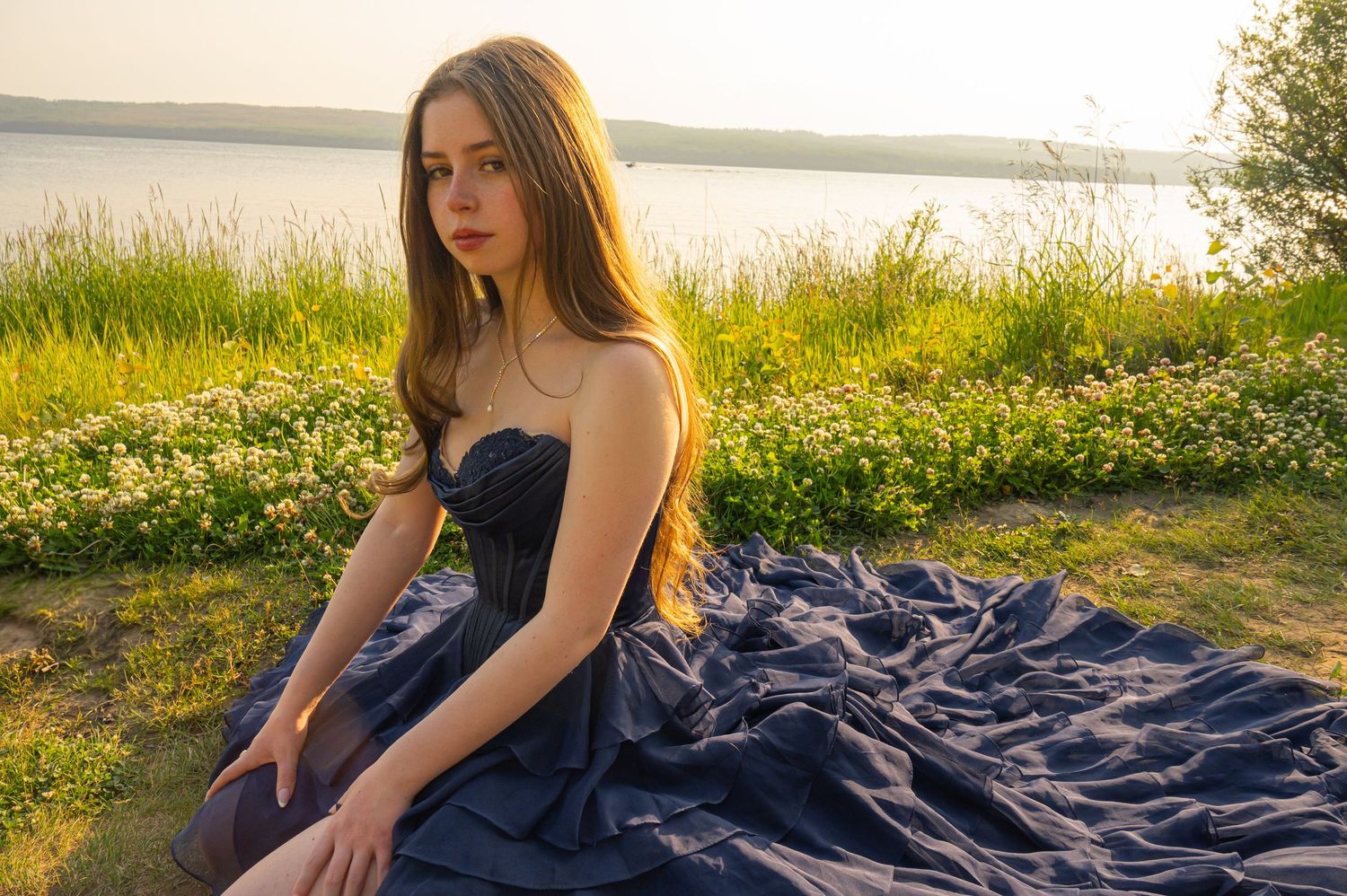 A person in a navy blue strapless gown poses near a lake at golden hour with wildflowers in the background.