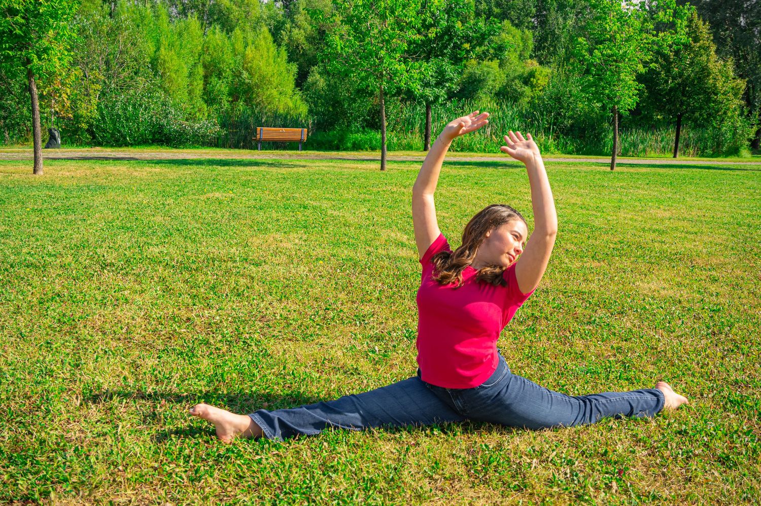 Person in a pink top performs a split stretch on green grass in a sunny park setting.