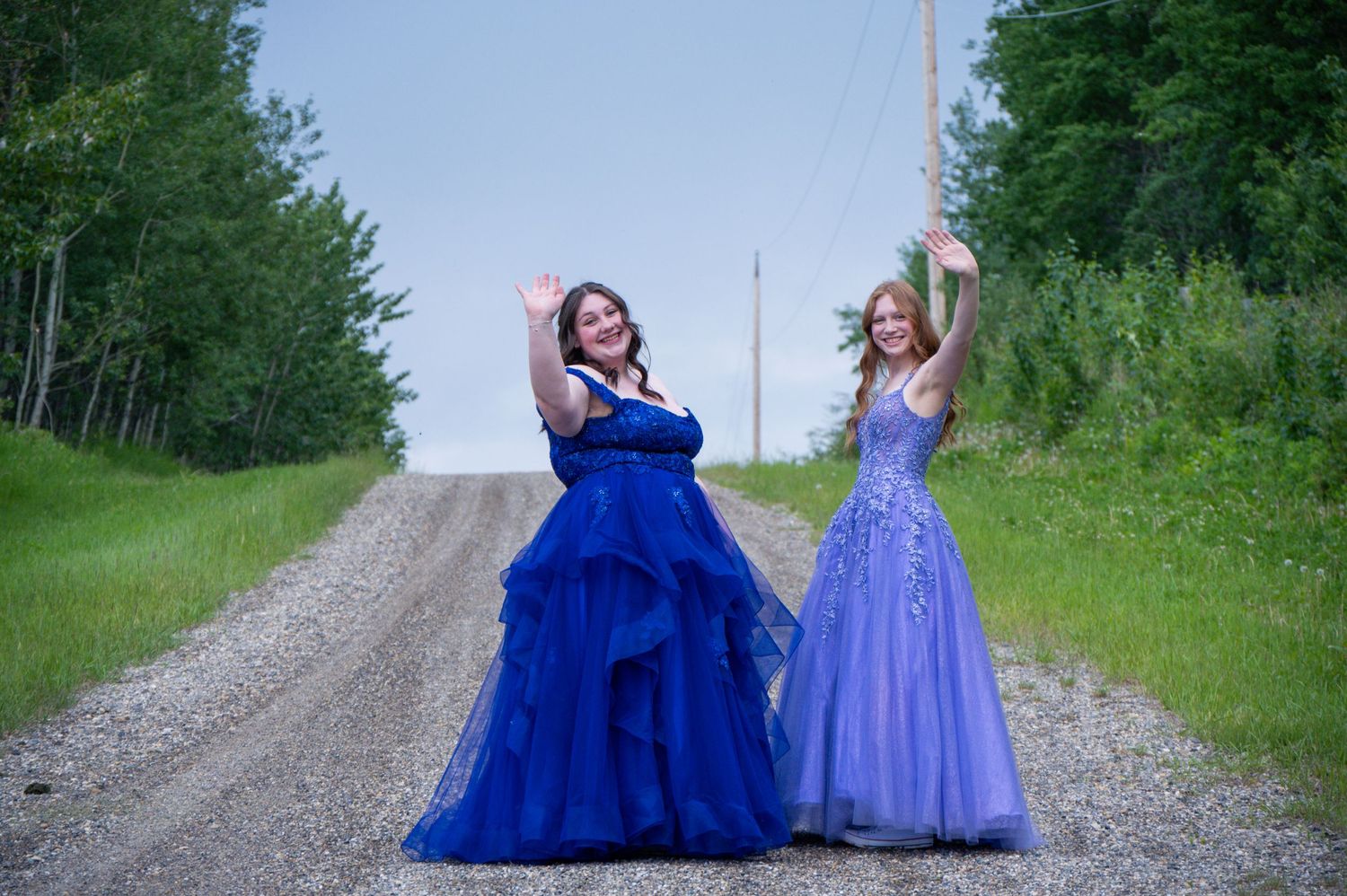 Two people in elegant formal gowns pose playfully on a rural dirt road surrounded by trees.