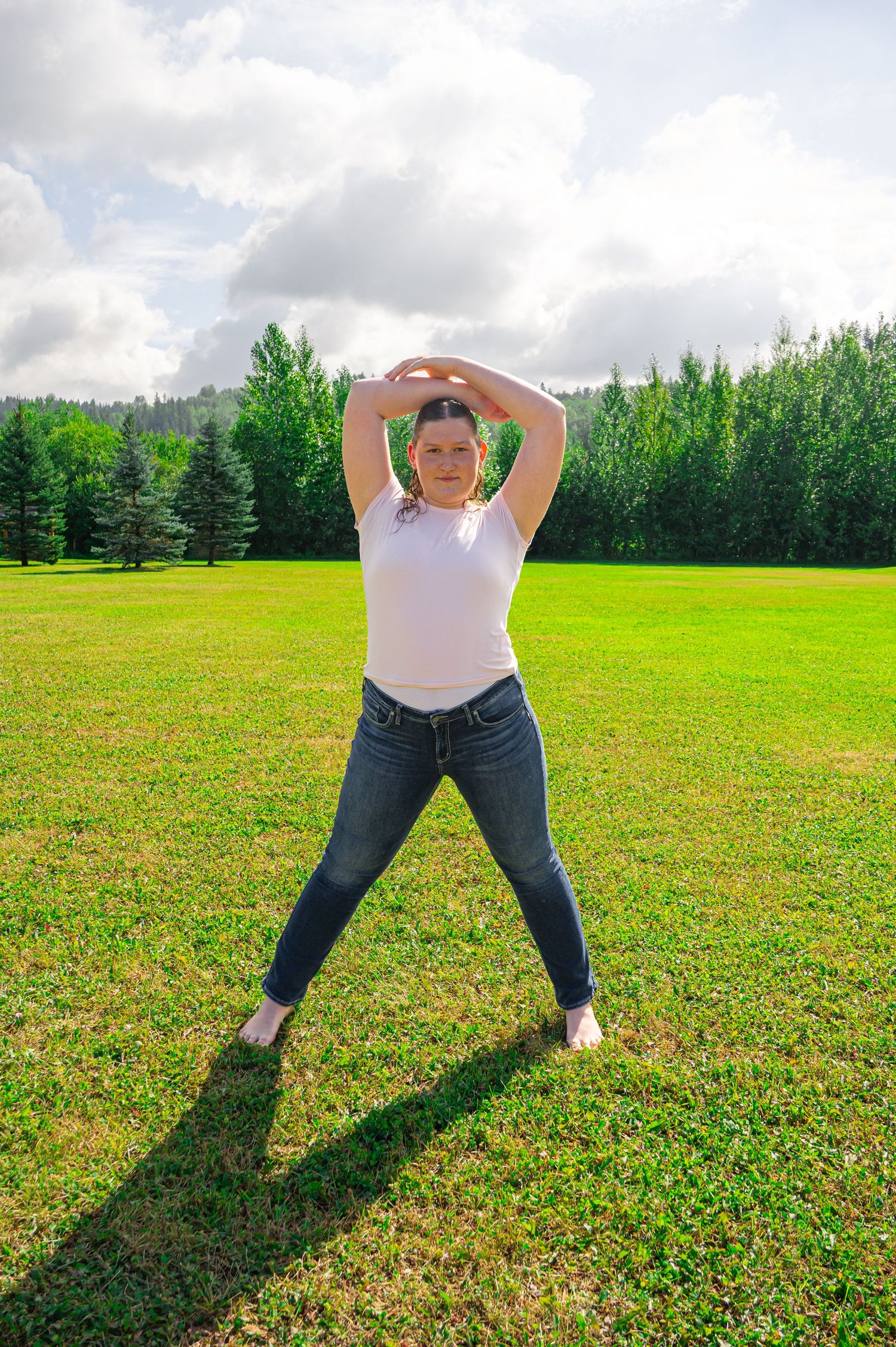 Someone strikes a playful pose in jeans and white top on a grassy field with forest backdrop.