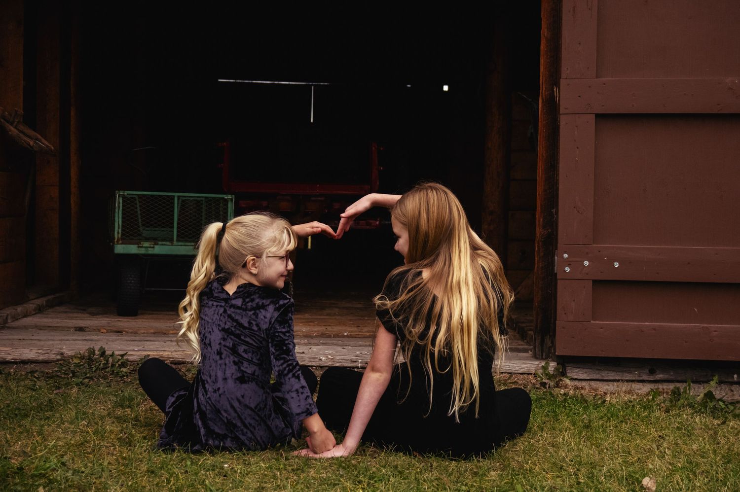 Two people sit casually on grass outside a rustic barn building.