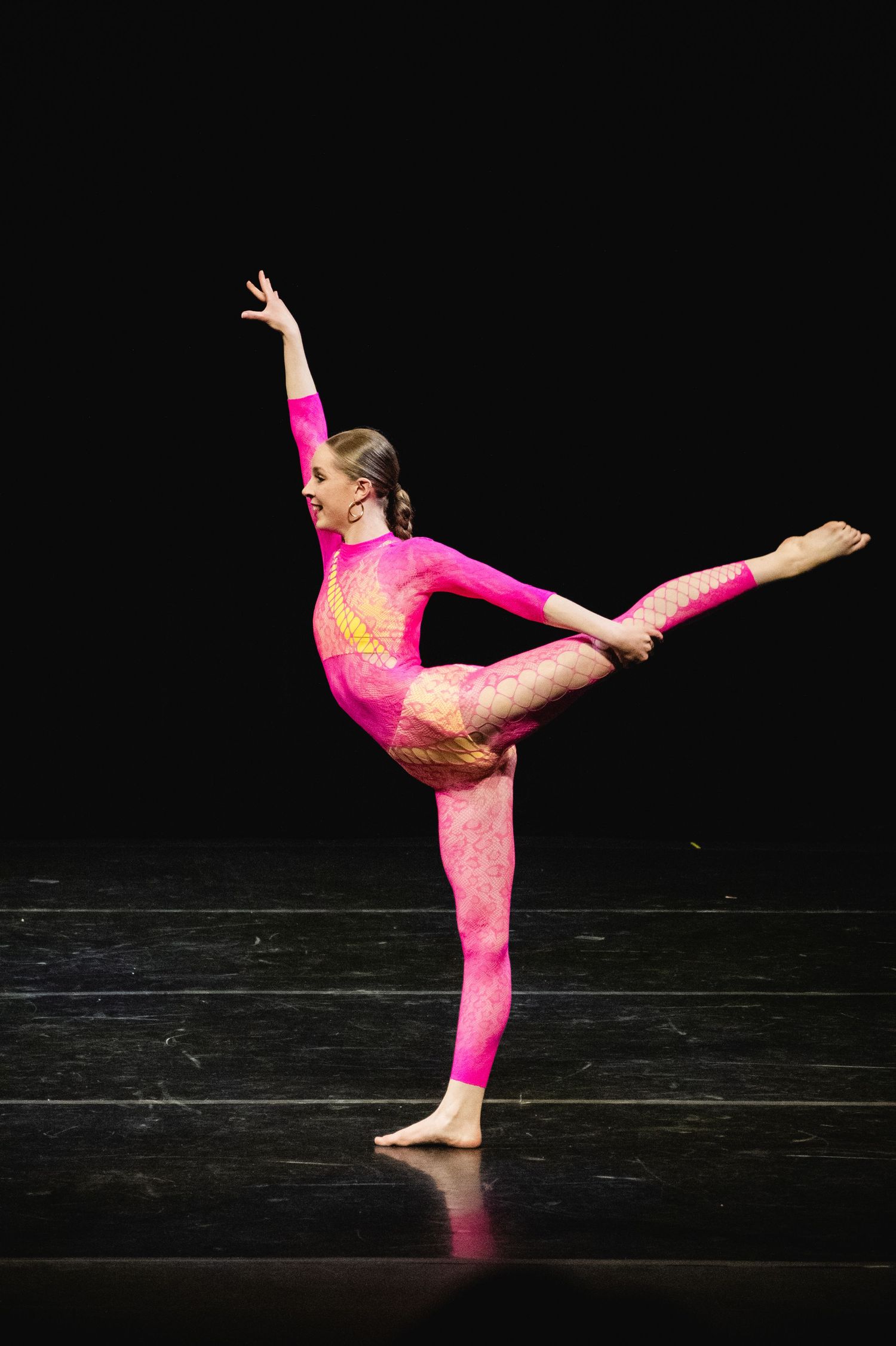 Dancer in bright pink leotard performs an artistic pose on dark stage, demonstrating graceful balance and flexibility.