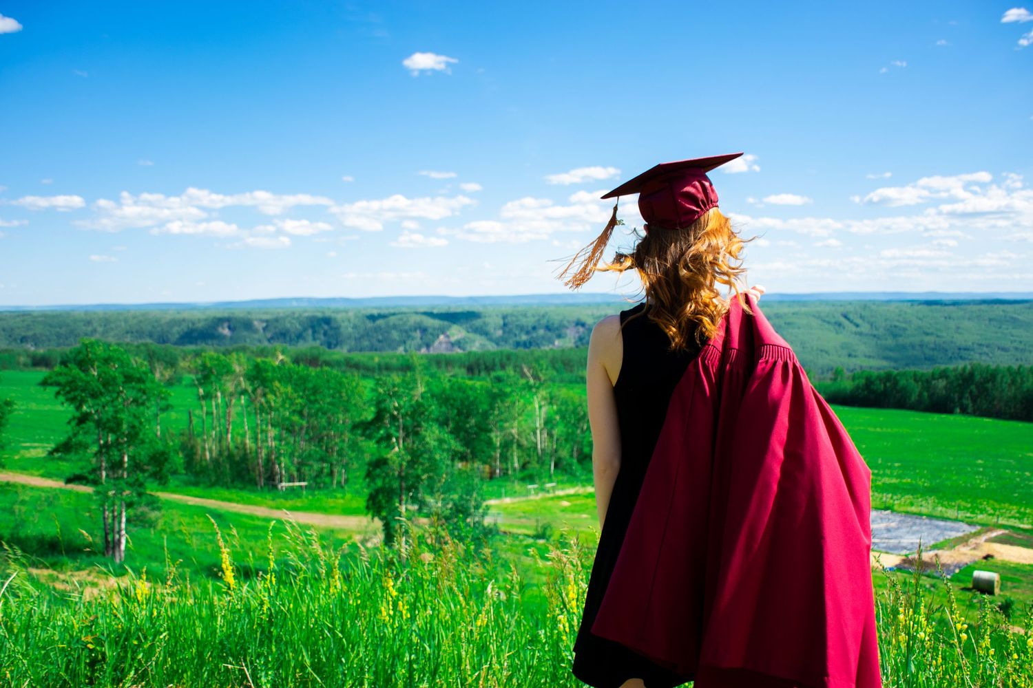 Graduate in red cap and gown looks out over rolling green hills.