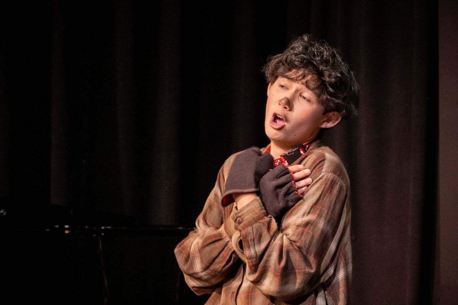A performer in a brown shirt gestures dramatically on stage during a theatrical performance as they sing or act.